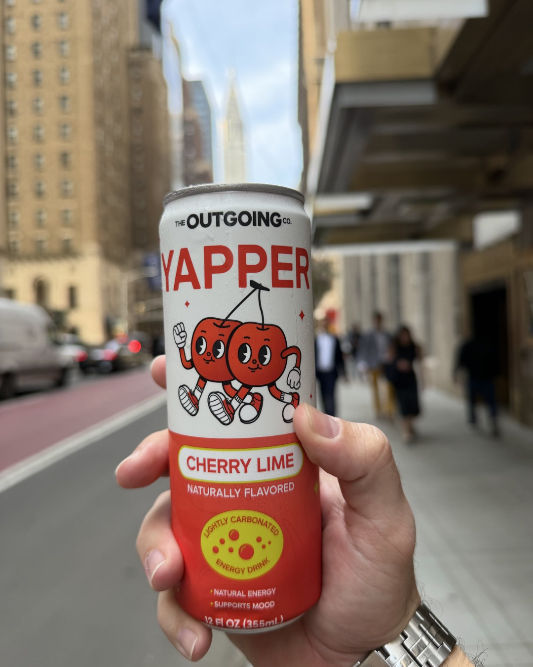 A hand holds a can of Yapper Cherry Lime energy drink on a city street.