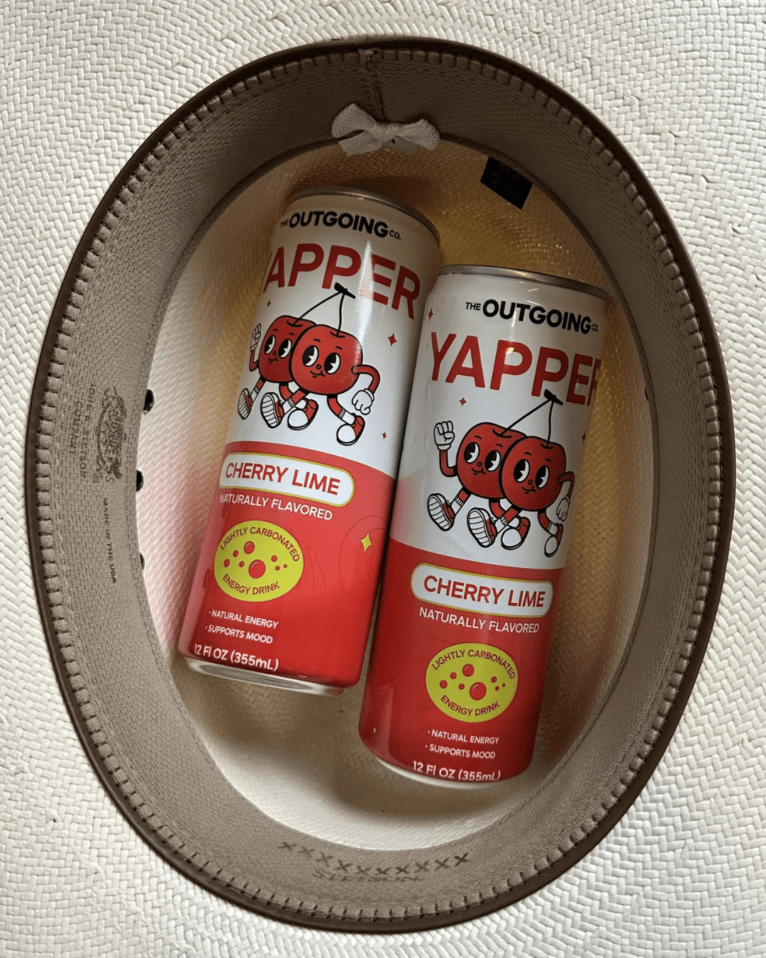 Two cans of The Outgoing Co. Yapper Cherry Lime energy drink sit inside a woven hat.