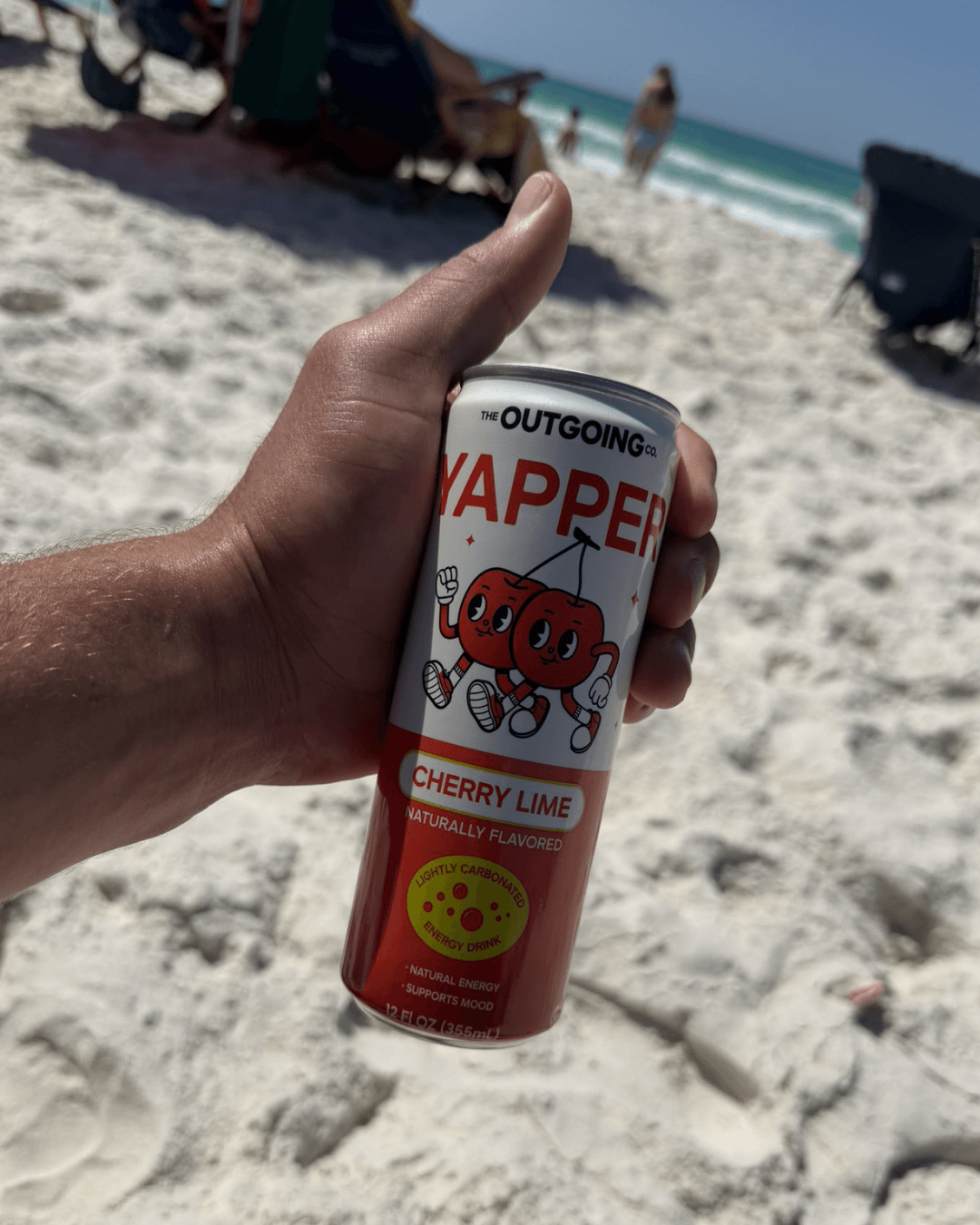 A hand holds a can of Yapper Cherry Lime energy drink on a white sand beach.