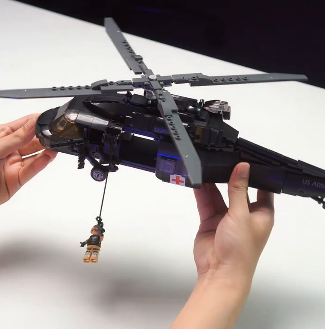 Hands hold a black Lego-style helicopter with a minifigure dangling from a rope beneath it.