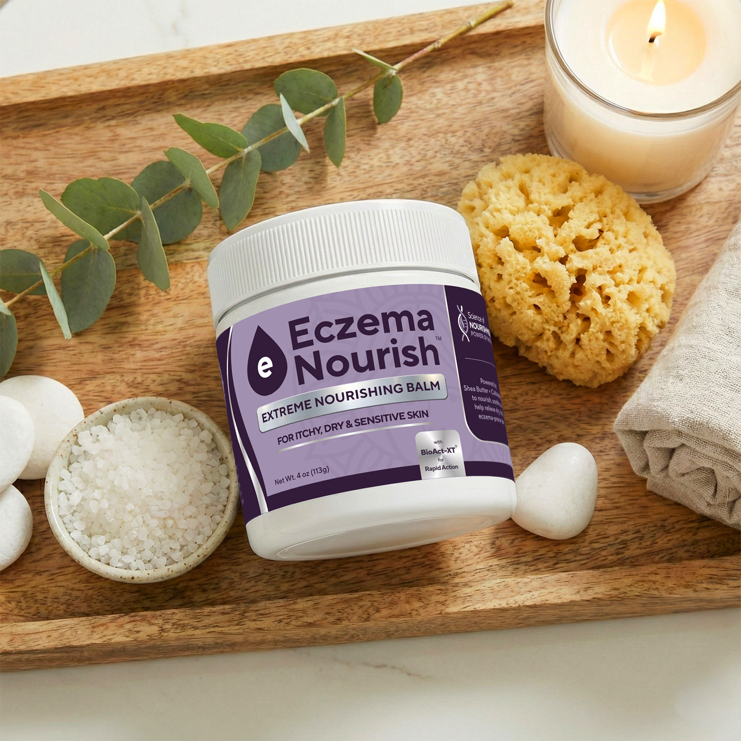 A jar of Eczema Nourish balm sits on a wooden tray with a candle, sponge, and bath salts.