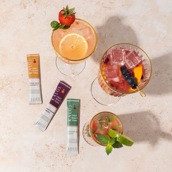 Three colorful drinks with fruit garnishes and tonic packets on a light surface.