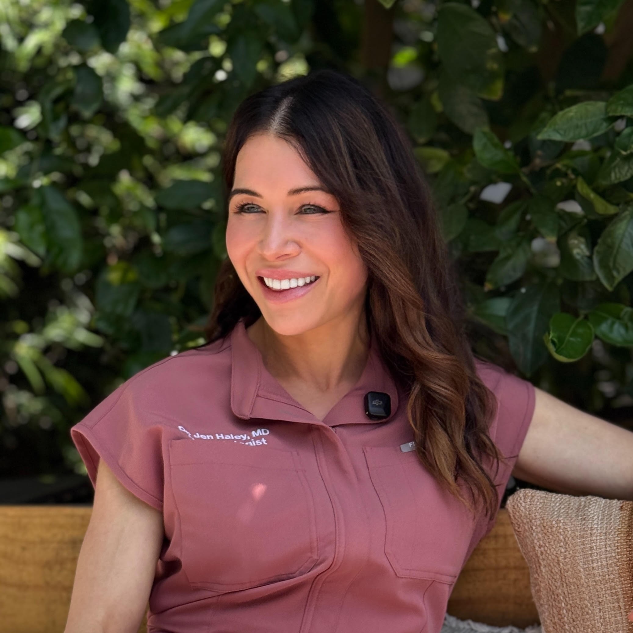 Smiling person in a pink shirt sitting outdoors with green foliage background.
