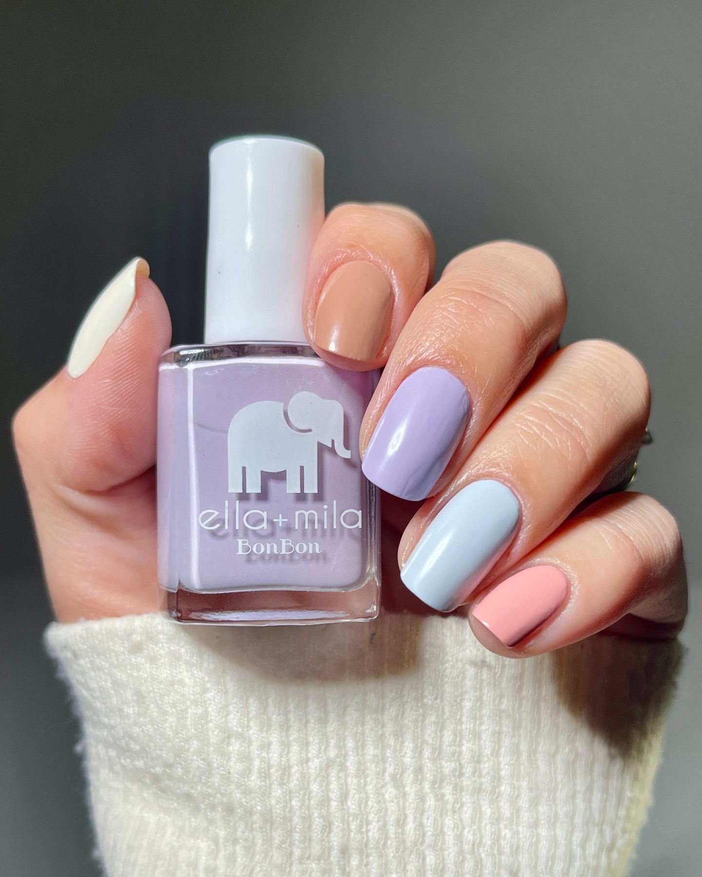 Hand holding a bottle of pastel purple nail polish with multicolored painted nails.
