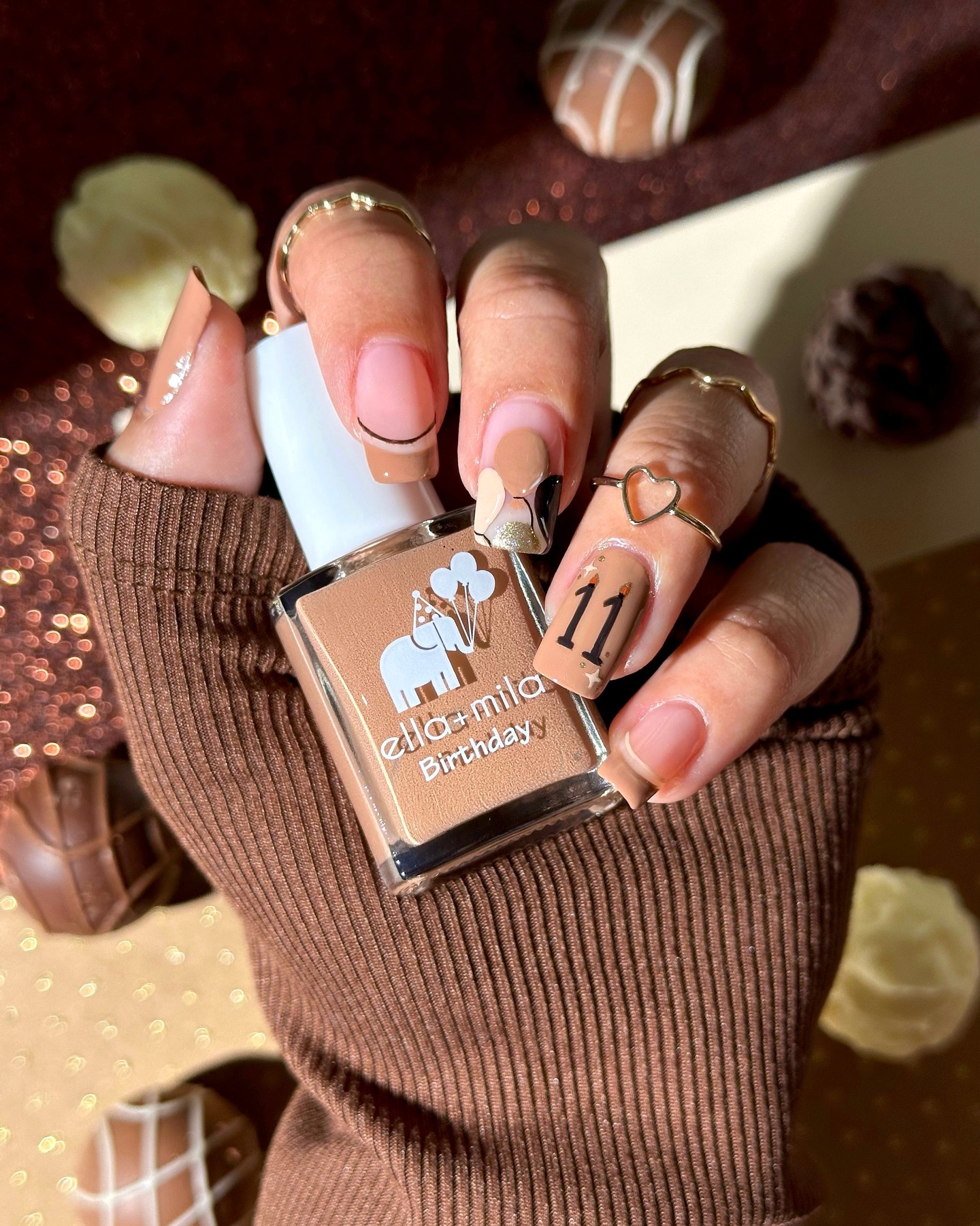 Hand holding nail polish bottle with decorated nails and chocolates in the background.