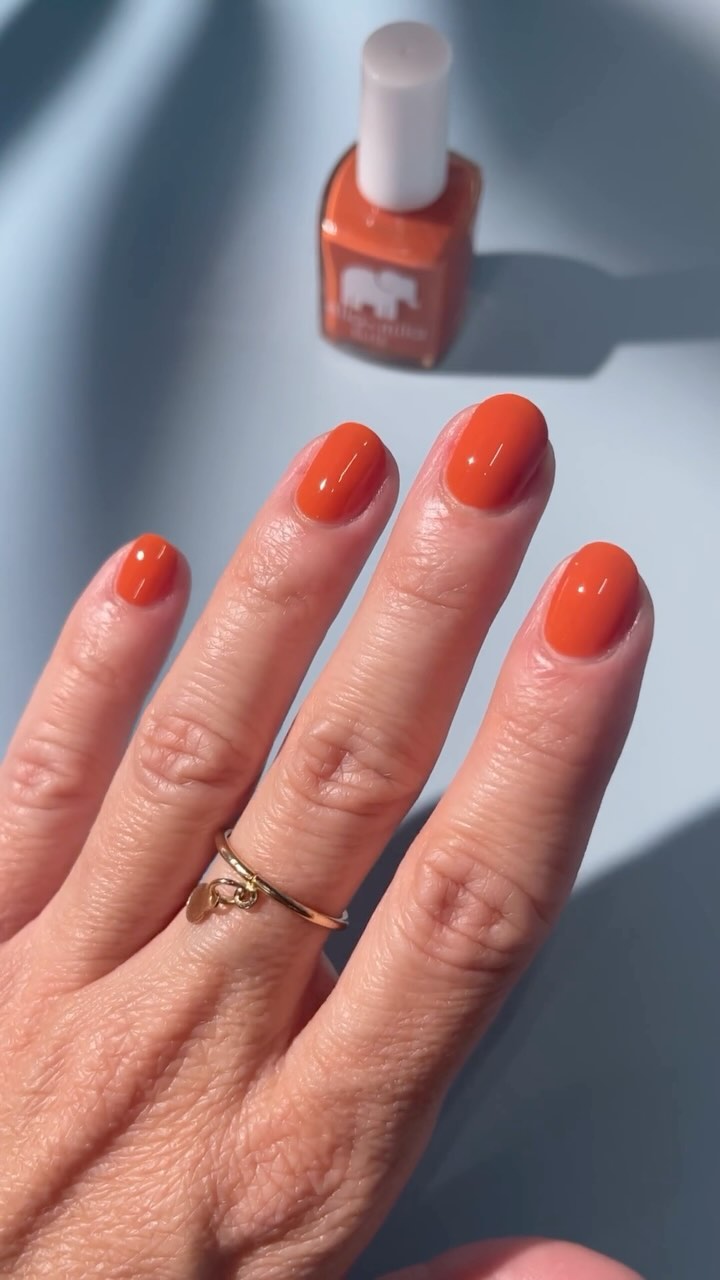 Hand with orange nail polish and a gold ring, nail polish bottle in background.