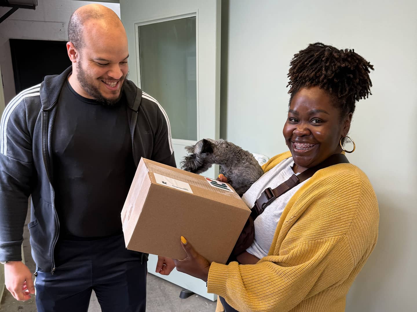 A smiling man looks at a cardboard box held by a smiling woman who is also holding a dog.