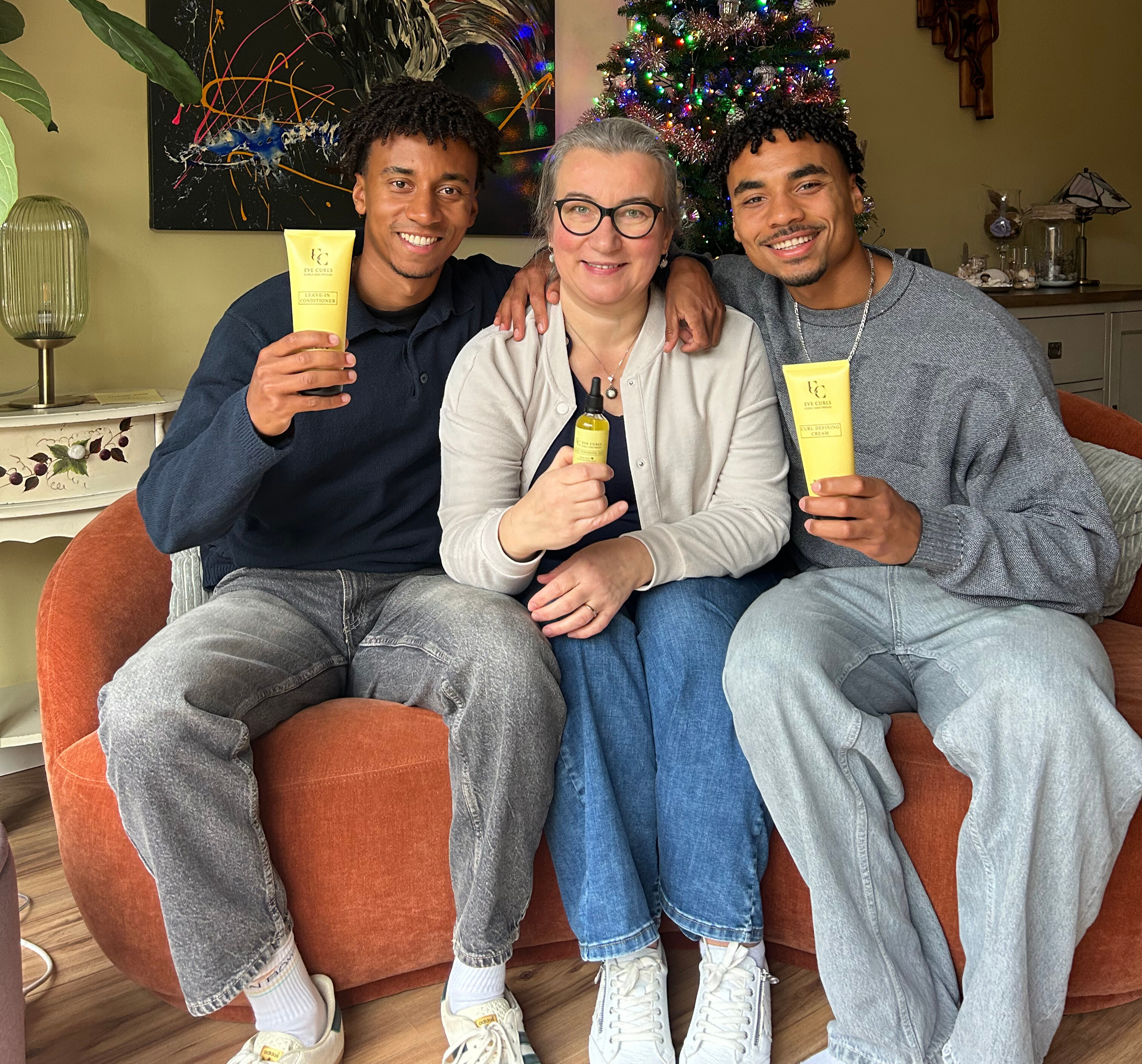 Three people sitting on a couch holding skincare products, with a decorated Christmas tree behind them.