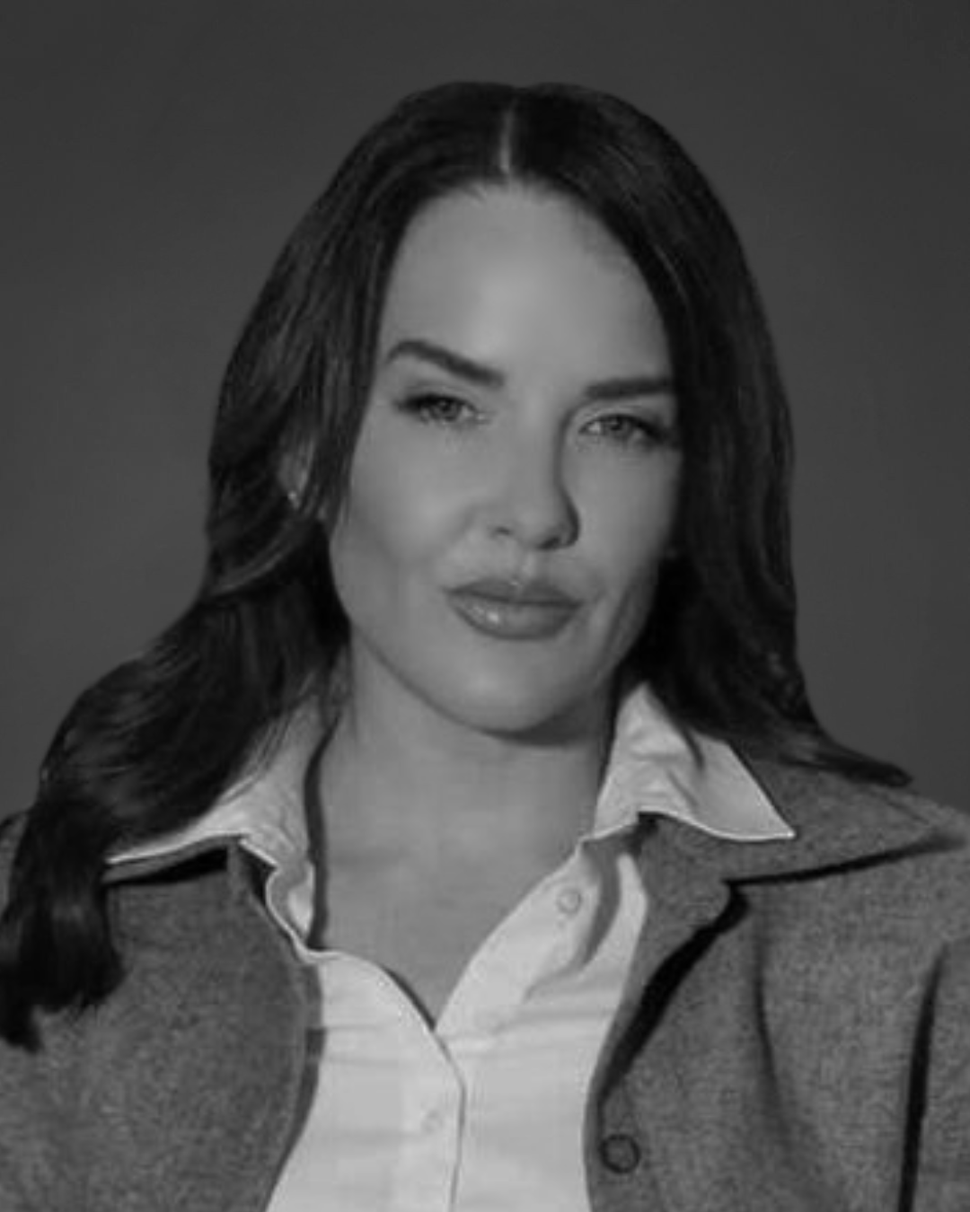 A black and white headshot of a woman with long dark hair, wearing a collared shirt and jacket.