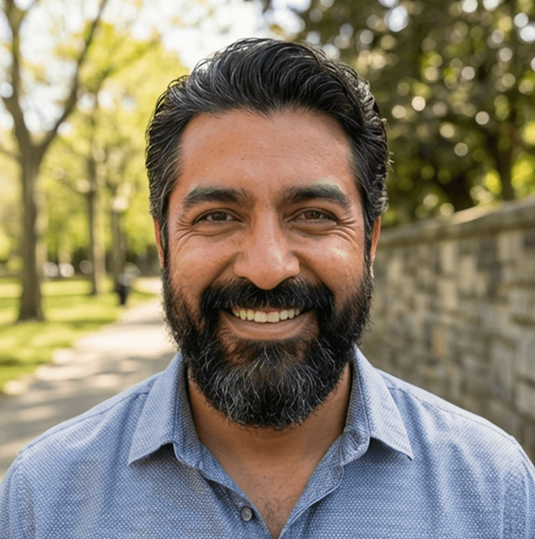 A headshot of a smiling man with a dark beard wearing a blue collared shirt outdoors.