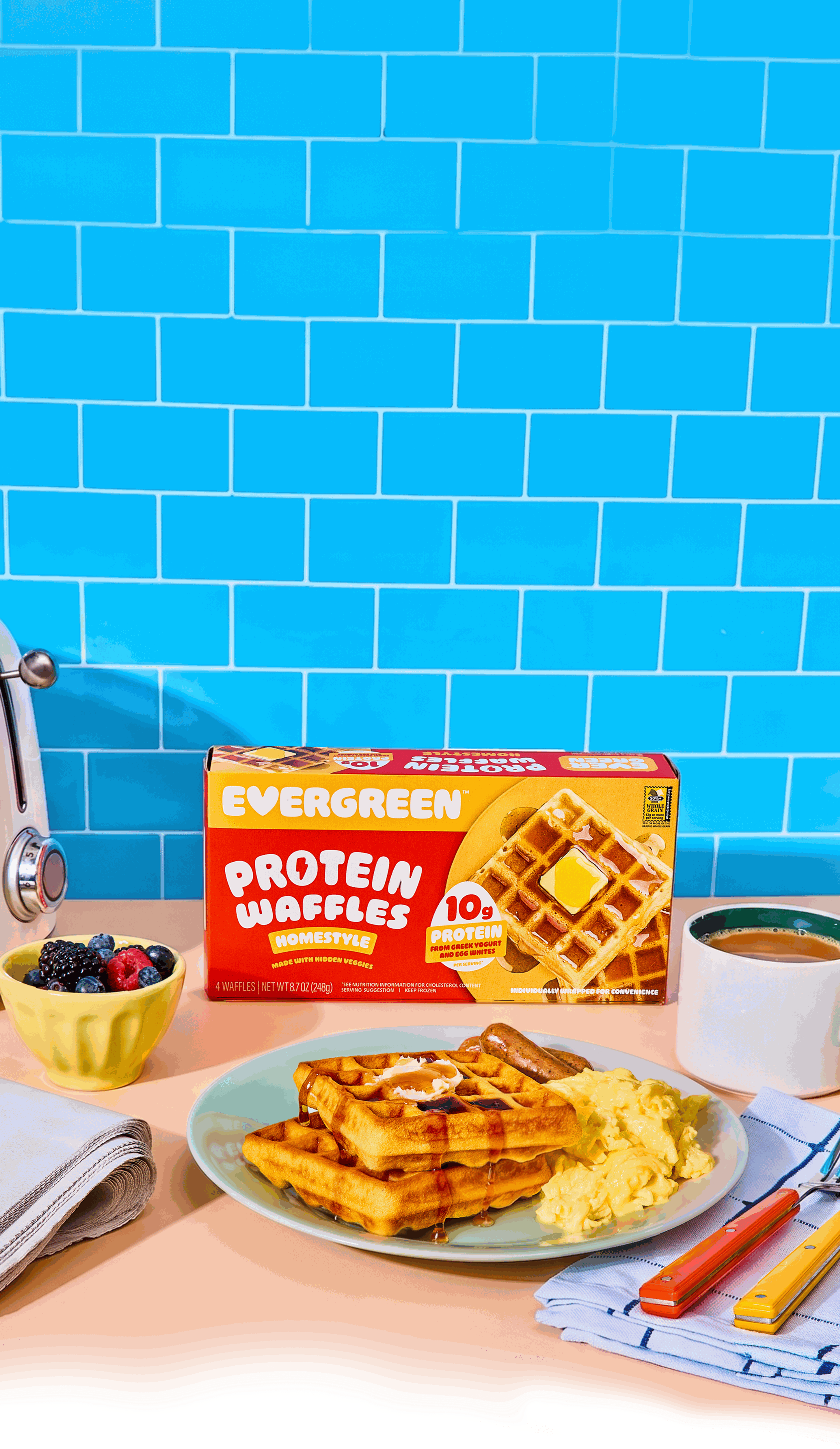 A box of Evergreen Protein Waffles sits behind a plated breakfast of waffles, eggs, and sausage.