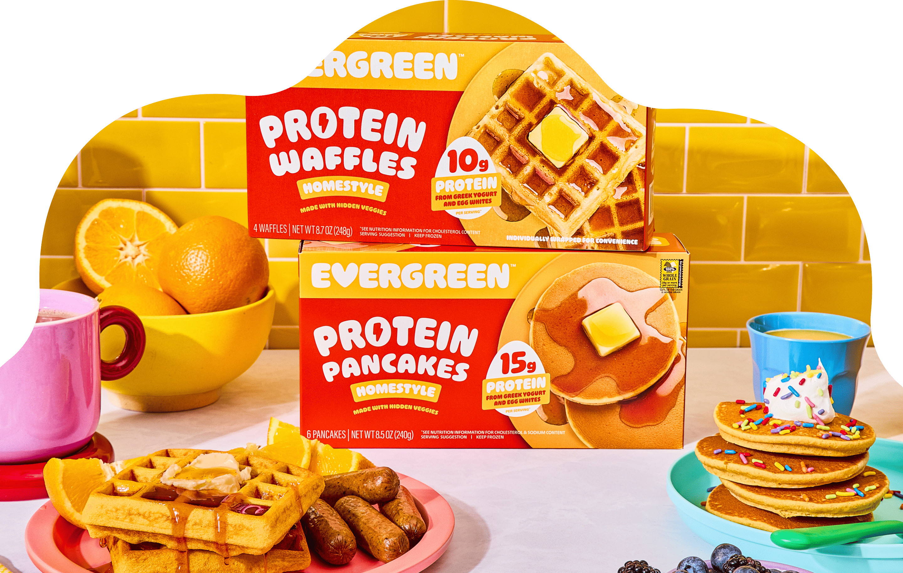 A colorful breakfast scene with boxes of Evergreen protein waffles and pancakes surrounded by plated breakfast foods.