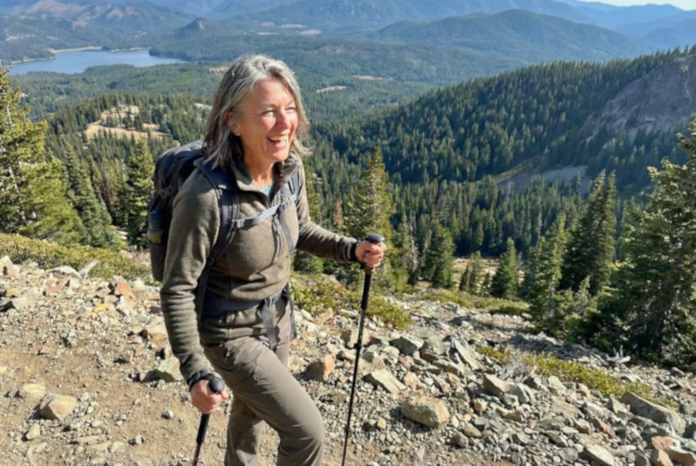 Smiling woman with backpack and trekking poles on a rocky alpine trail overlooking forested mountains and a lake