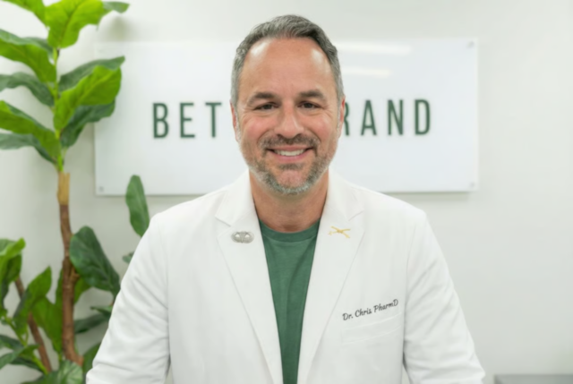 Dr. Chris PharmD smiling in white lab coat, standing before a sign reading 'BETRAND' with potted plants nearby