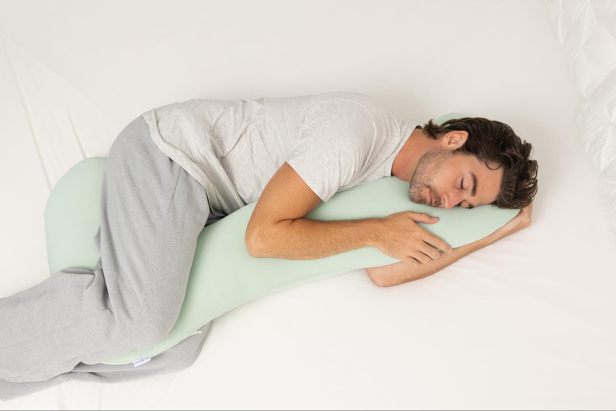 A man in pajamas sleeps on his side, hugging a large, light-green body pillow on a white bed.