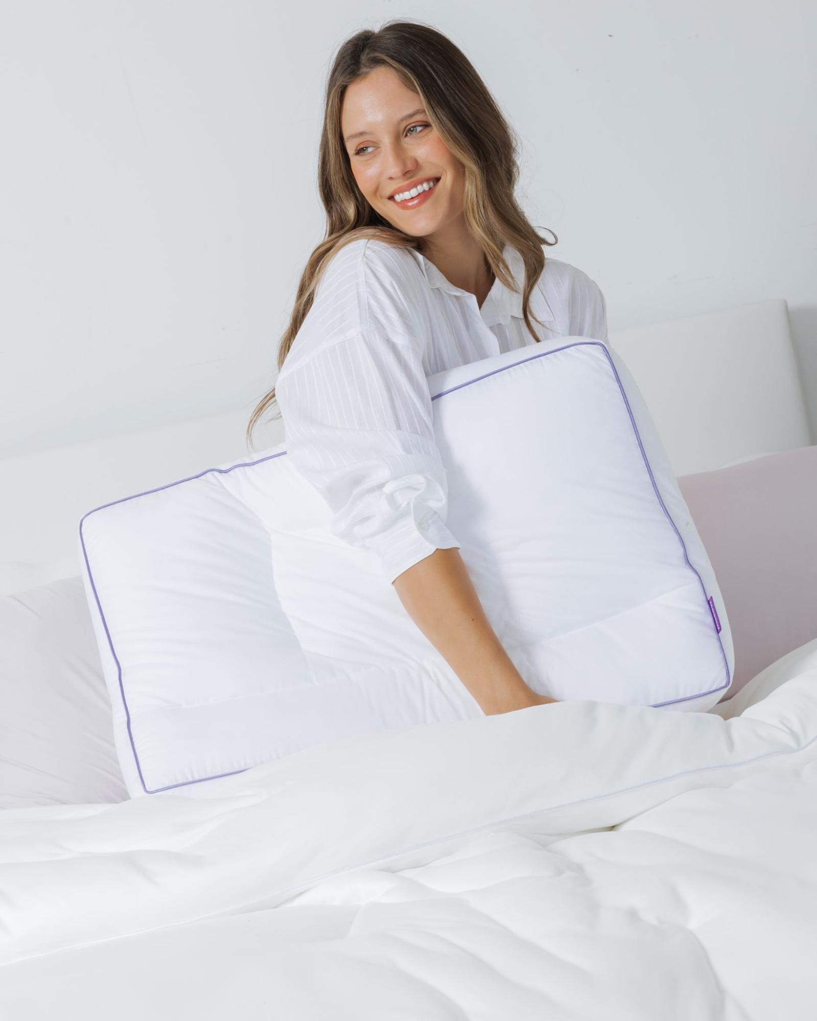 A smiling woman in a white shirt sits in bed, holding a white pillow with purple trim.