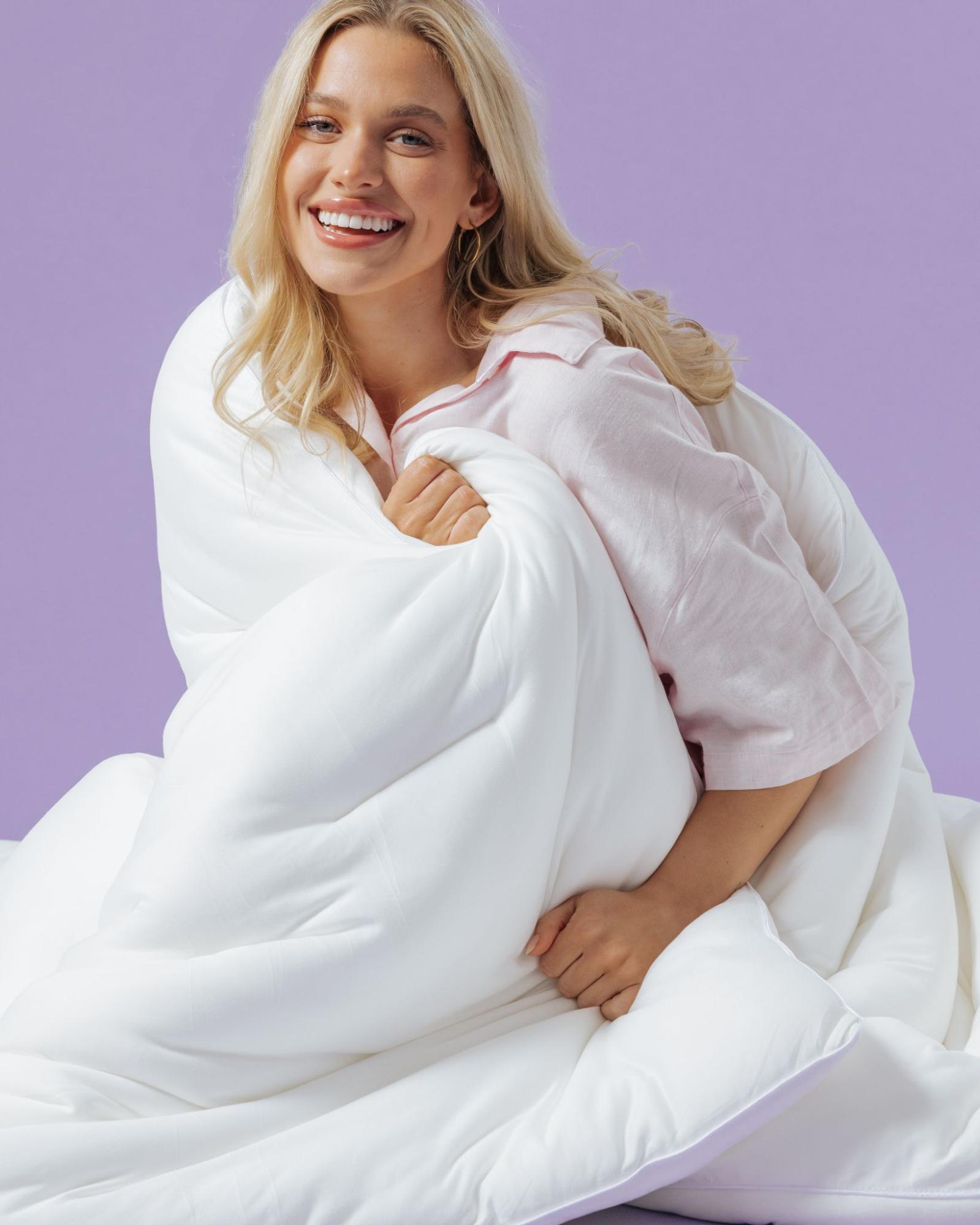 A smiling blonde woman in pink pajamas hugs a fluffy white comforter against a purple background.