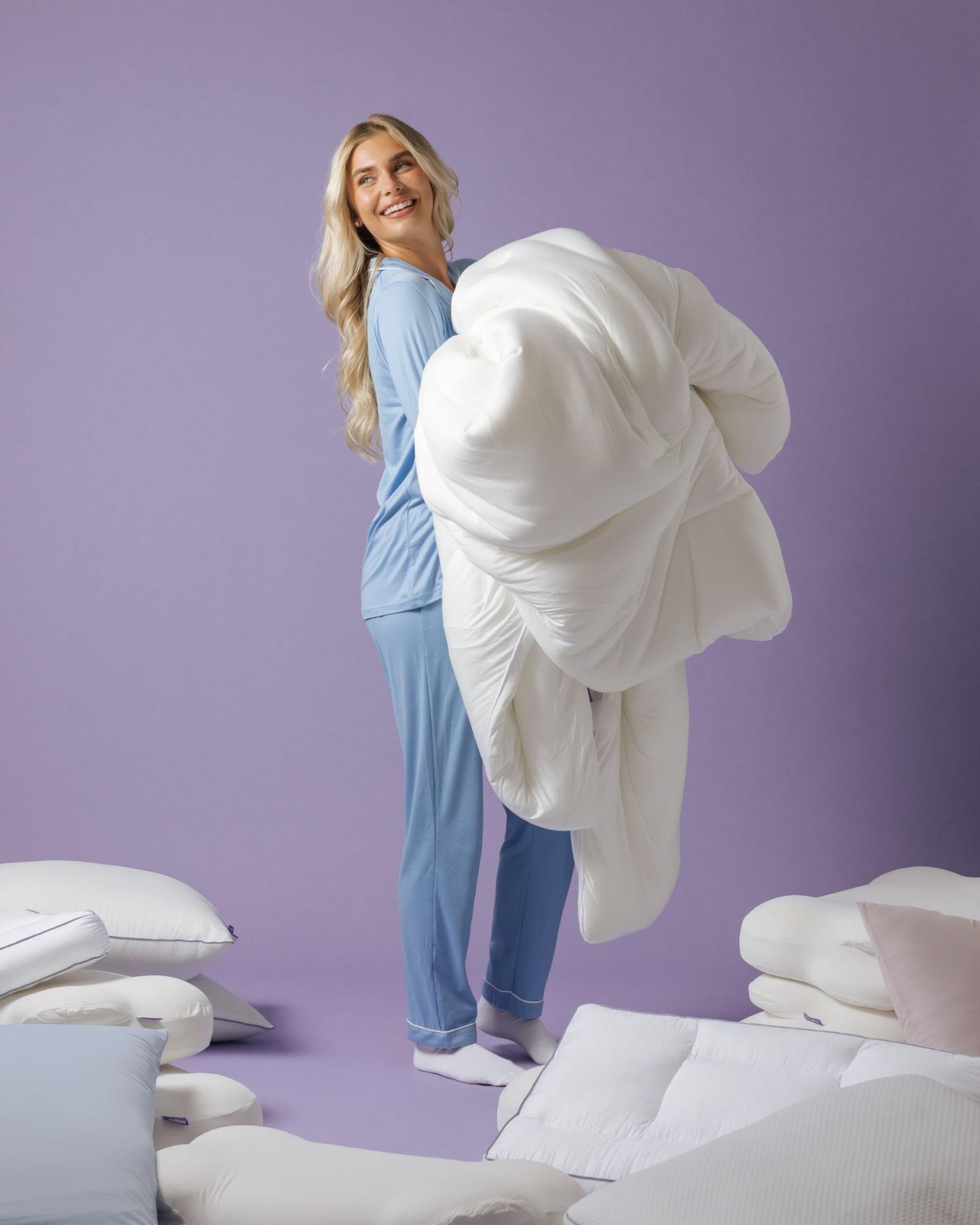 A smiling woman in blue pajamas holds a large white comforter while standing among many pillows.