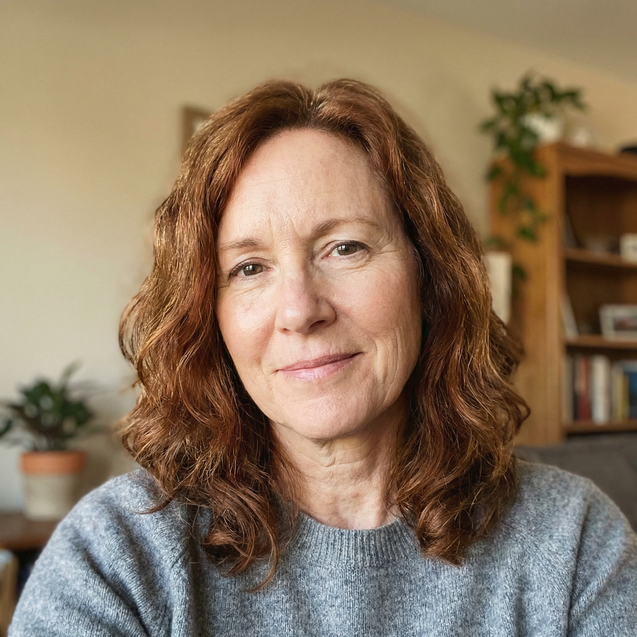A smiling woman with wavy reddish-brown hair, wearing a gray sweater, looks at the camera.