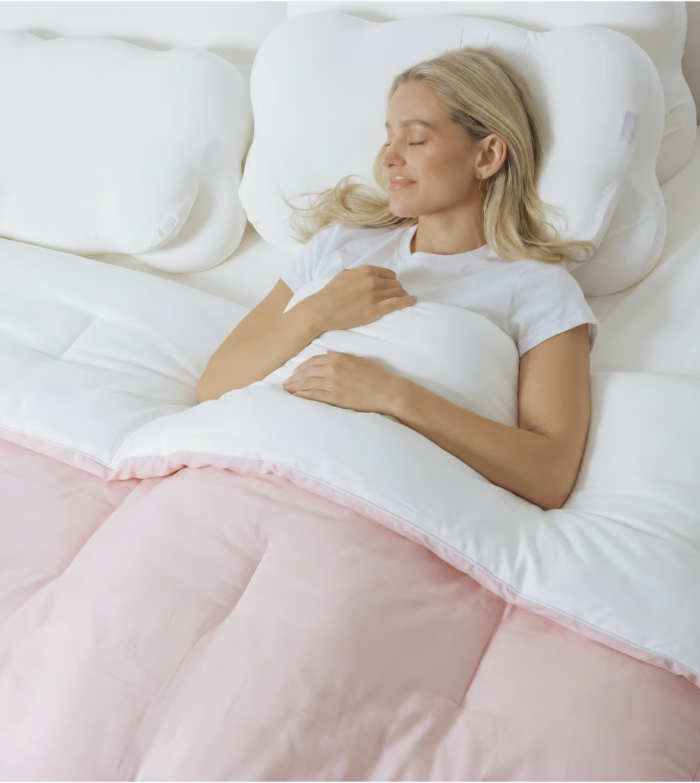 An overhead shot of a blonde woman sleeping peacefully in bed under a white and pink comforter.