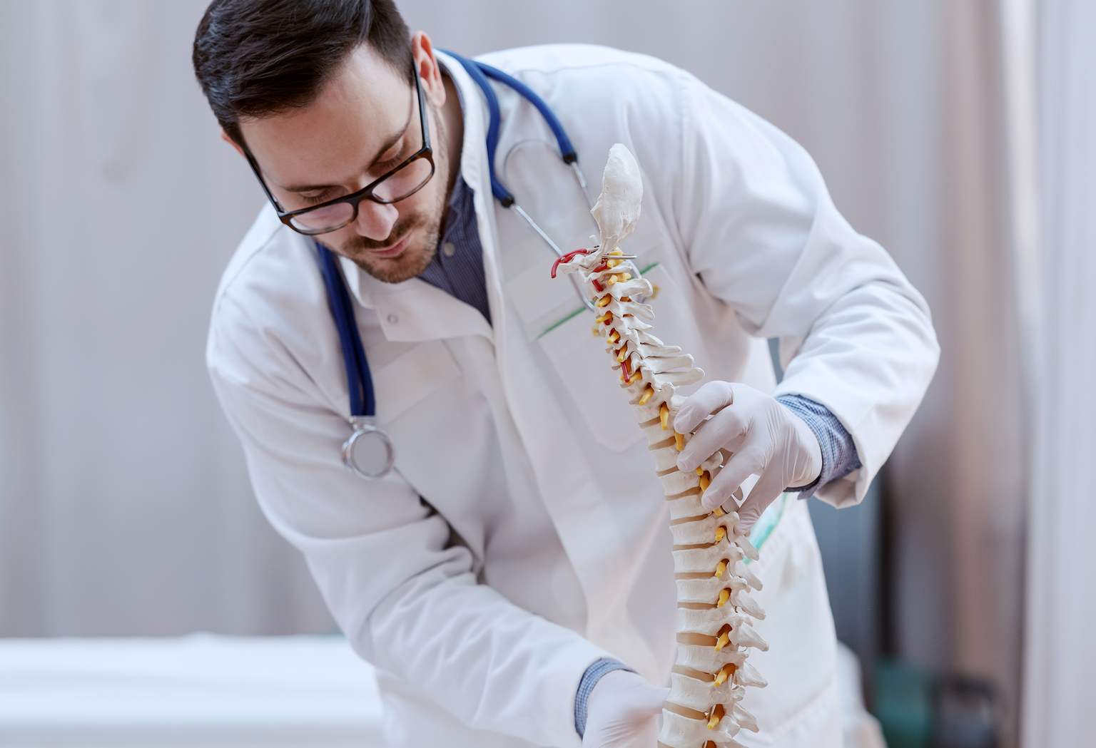A doctor in a white coat and glasses examines an anatomical model of a human spine.