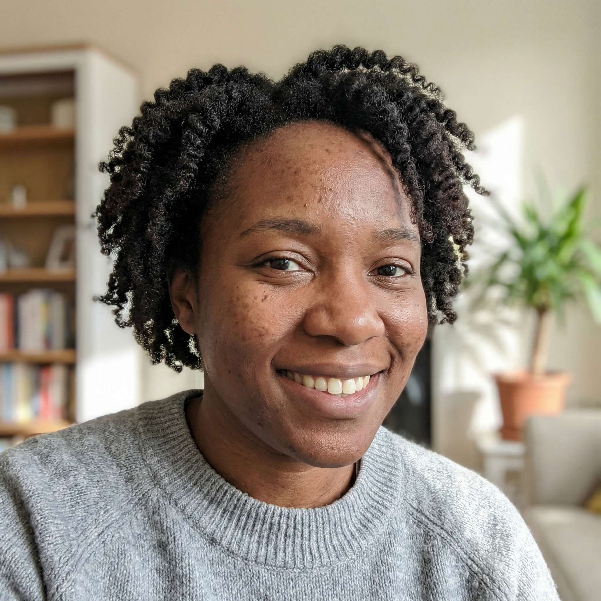 A smiling Black woman with short, curly hair, wearing a grey sweater in a sunlit room.