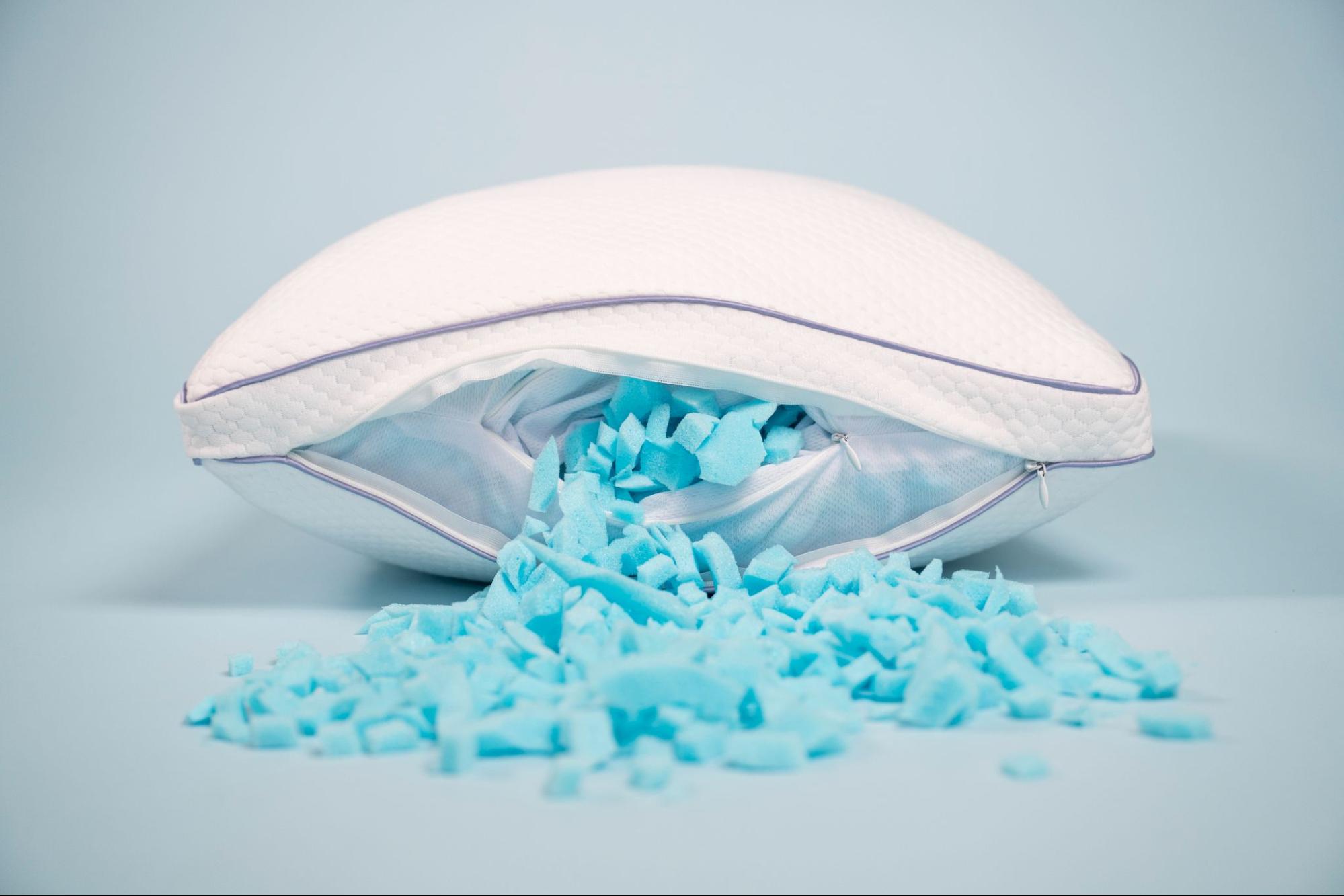A white pillow is unzipped, with its blue shredded foam filling spilling out onto a light blue surface.
