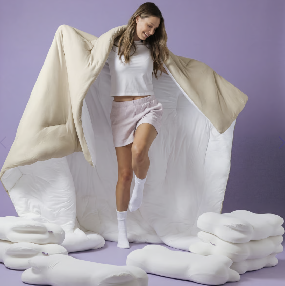 A smiling woman holds a large beige and white comforter around herself, surrounded by white pillows.