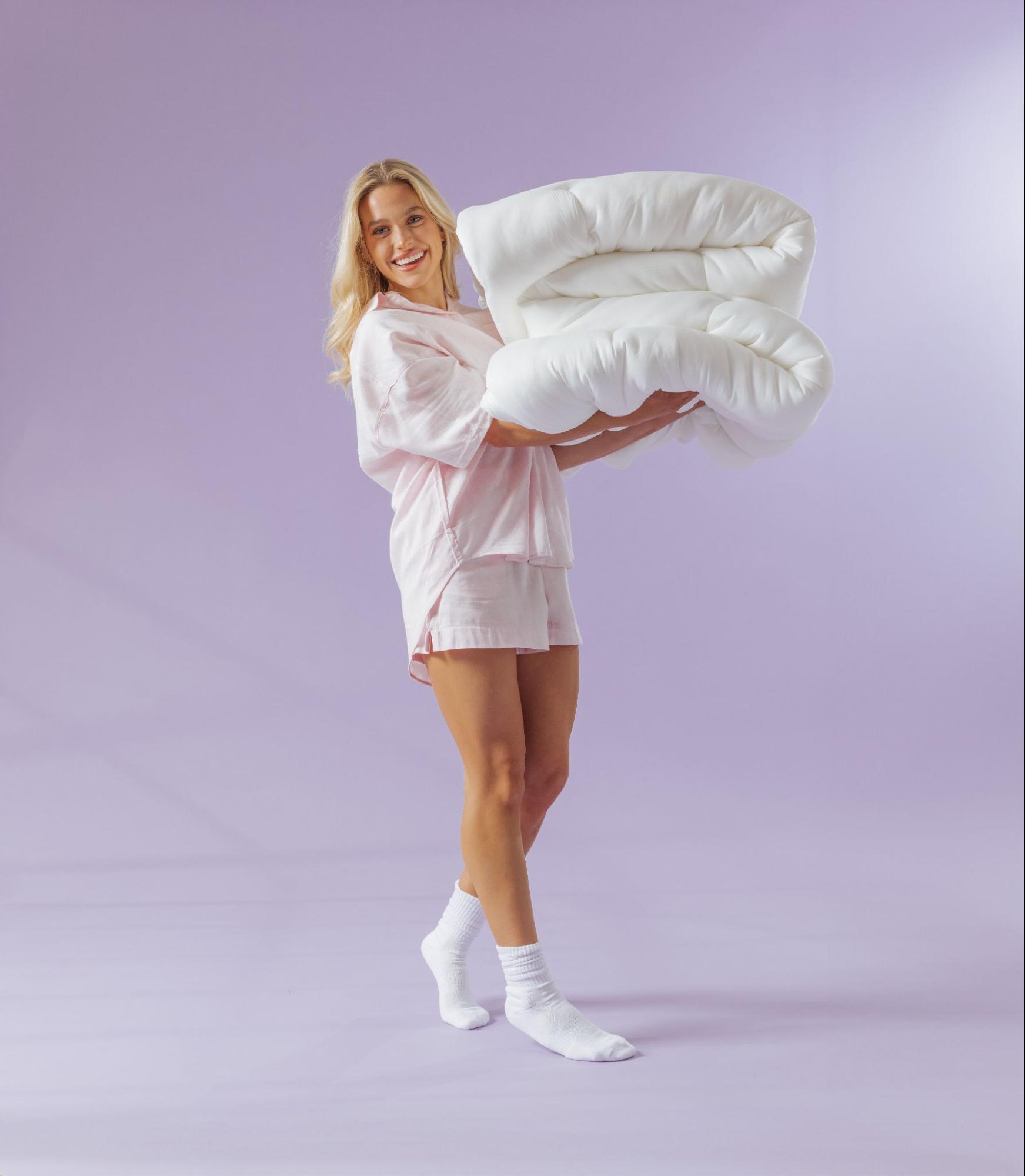 A smiling woman in pink pajamas and white socks holds a large, folded white comforter against a purple background.