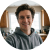 A circular, slightly blurry headshot of a smiling young man in a grey hoodie, indoors.