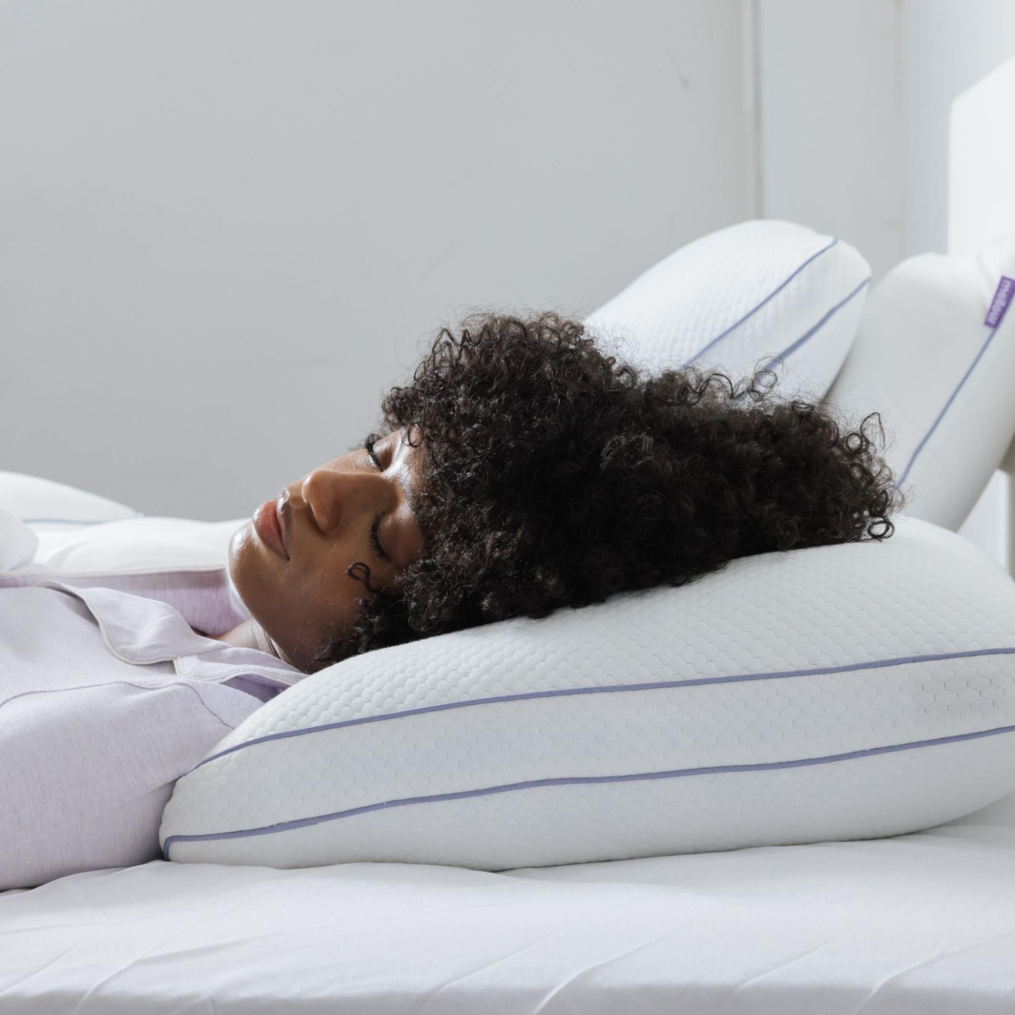A woman with dark curly hair sleeps on her back on a contoured white pillow with purple piping.