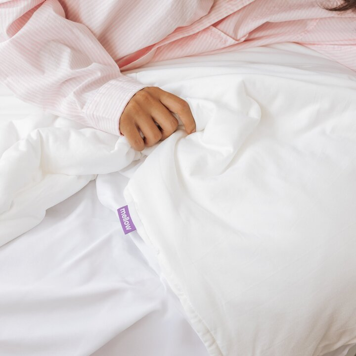 A person wearing pink striped pajamas holds a white comforter with a purple tag that says 'mellow'.