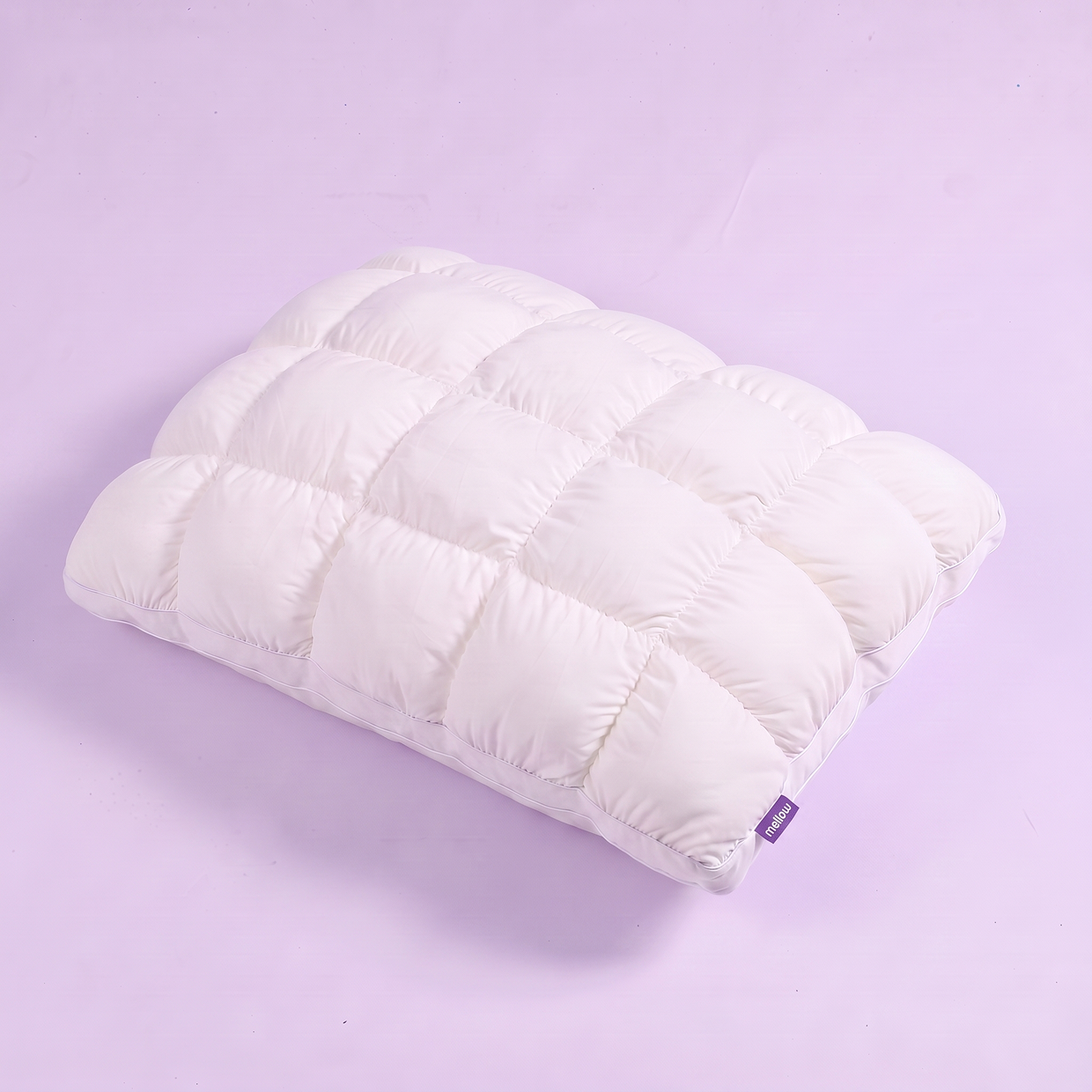 A white quilted pillow with a small purple brand tag, shown at an angle on a lavender background.