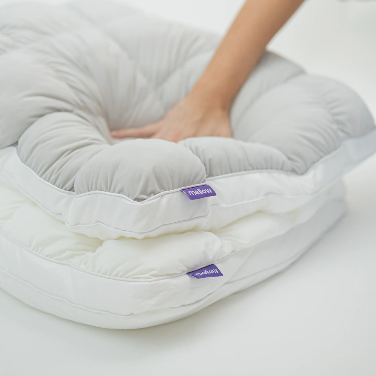 A hand presses down on a plush, two-toned grey and white Mellow mattress topper against a white background.