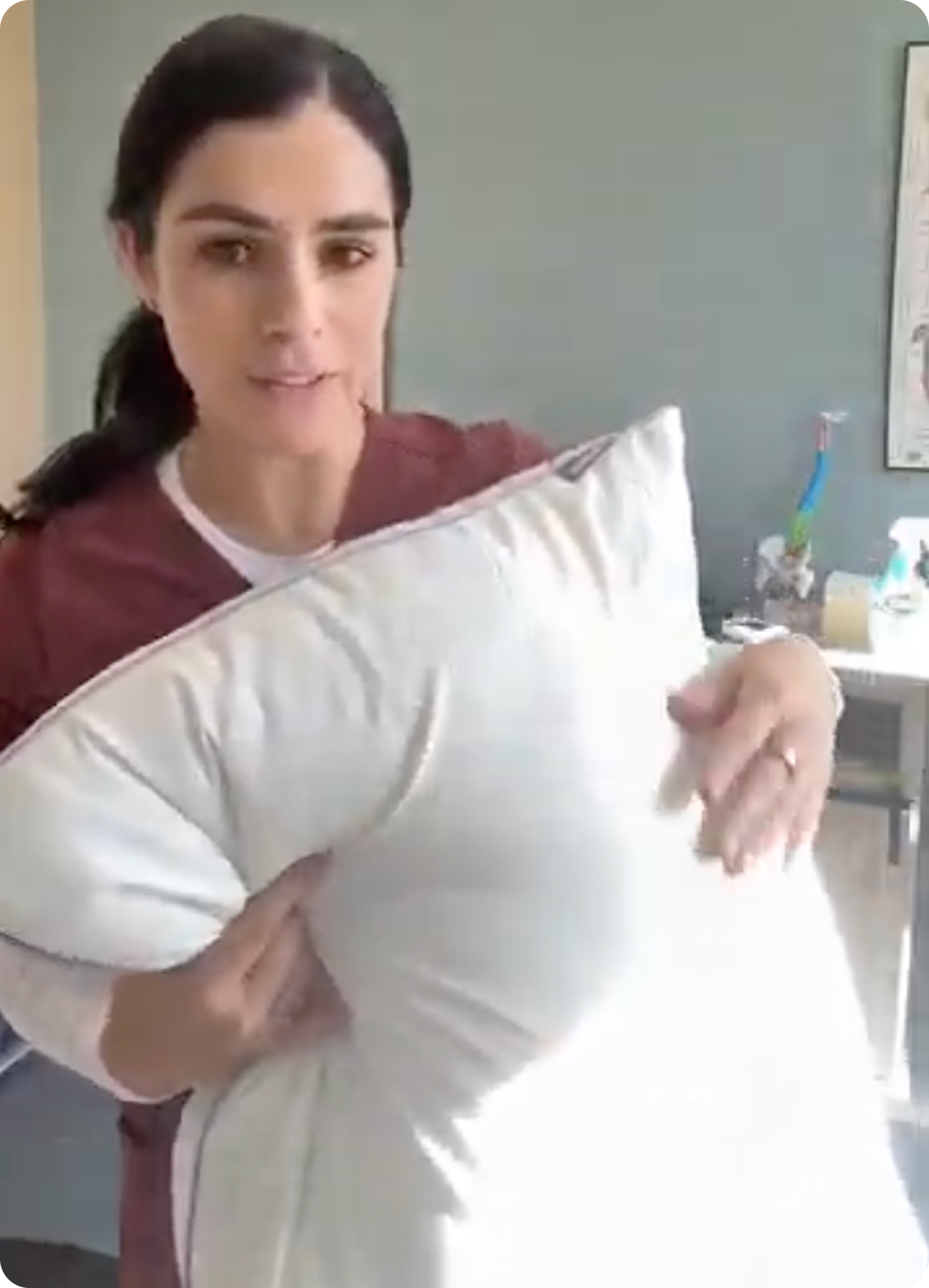 A woman with dark hair, wearing a reddish-brown top, holds a large white pillow and looks forward.
