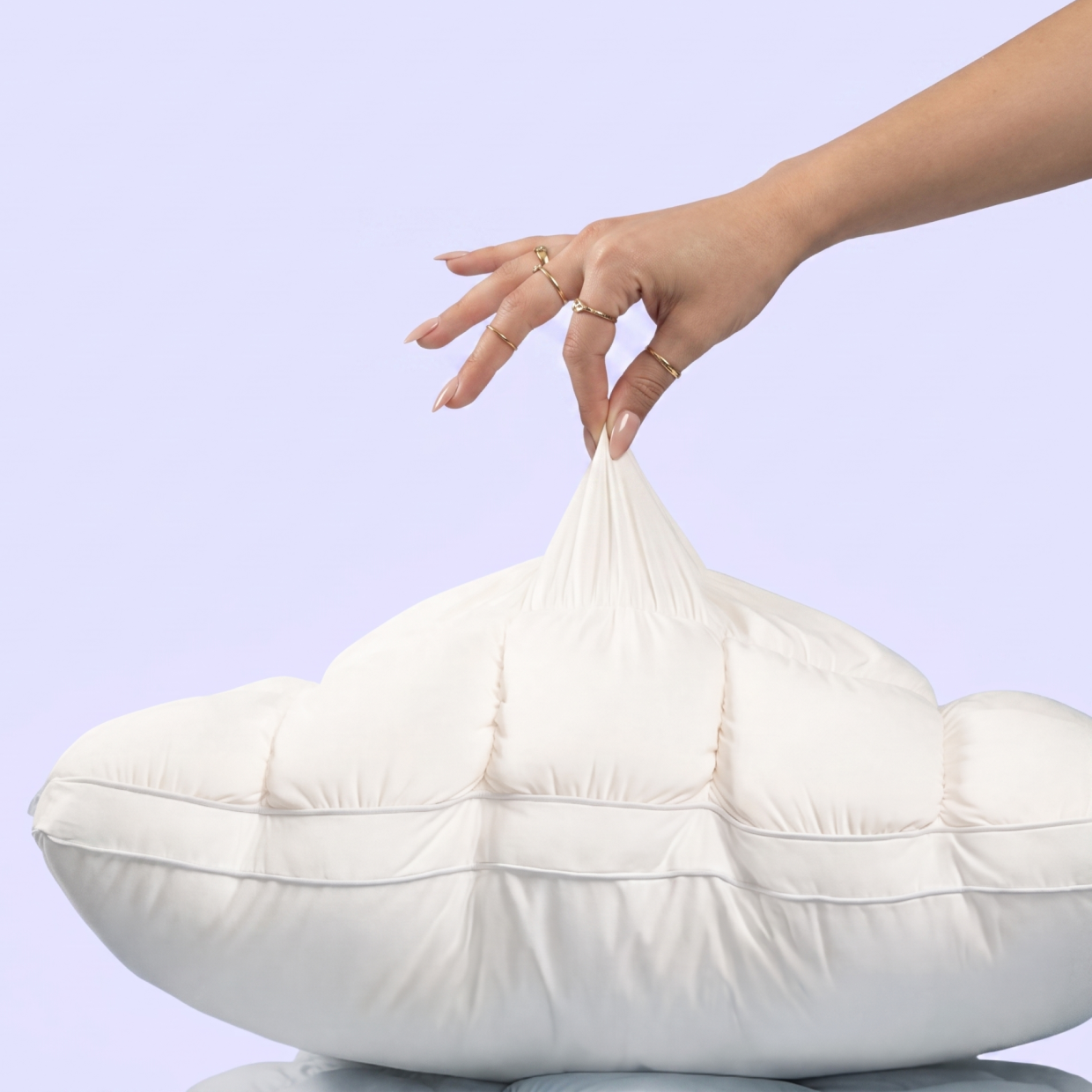 A hand wearing gold rings pinches the fabric of a white quilted pillow against a light purple background.