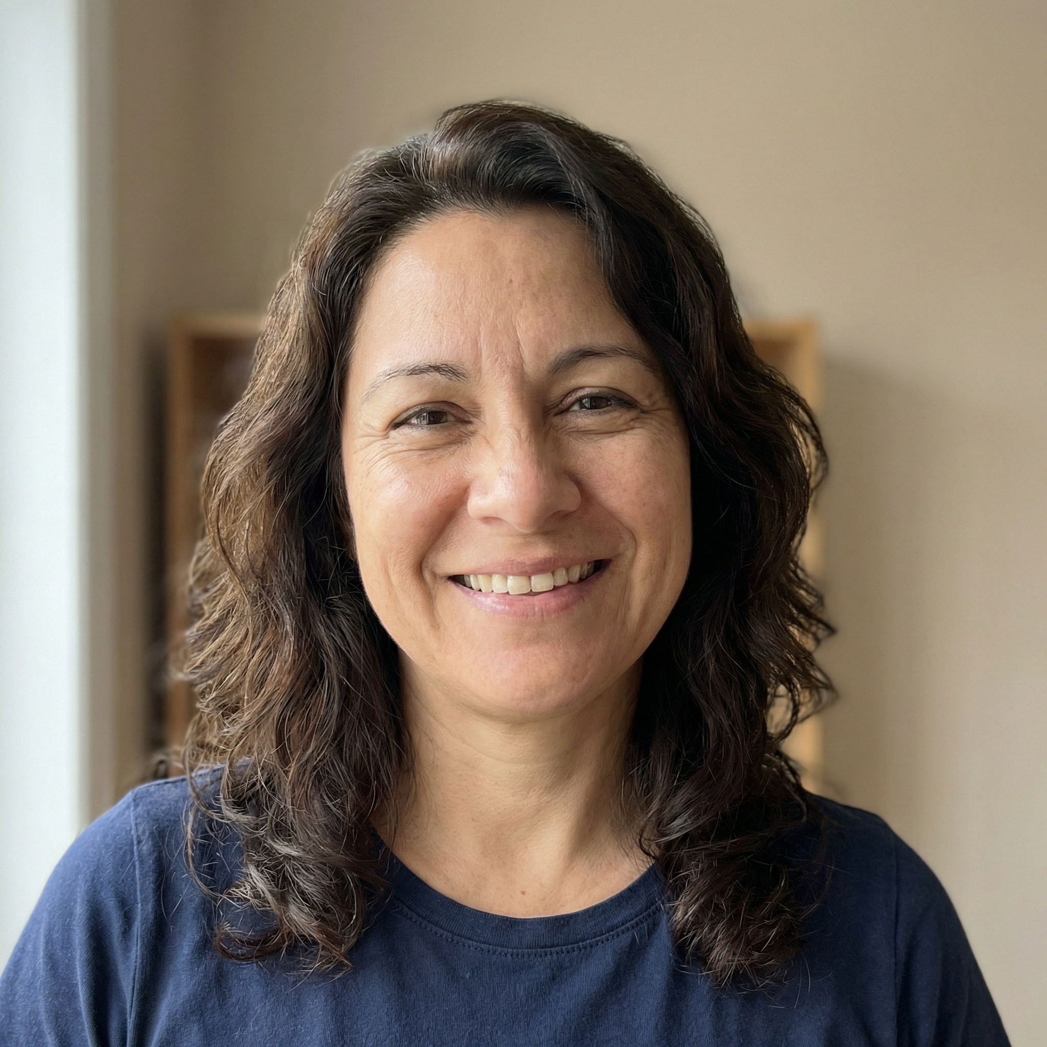 A headshot of a smiling woman with dark, wavy hair, wearing a blue shirt against a neutral background.