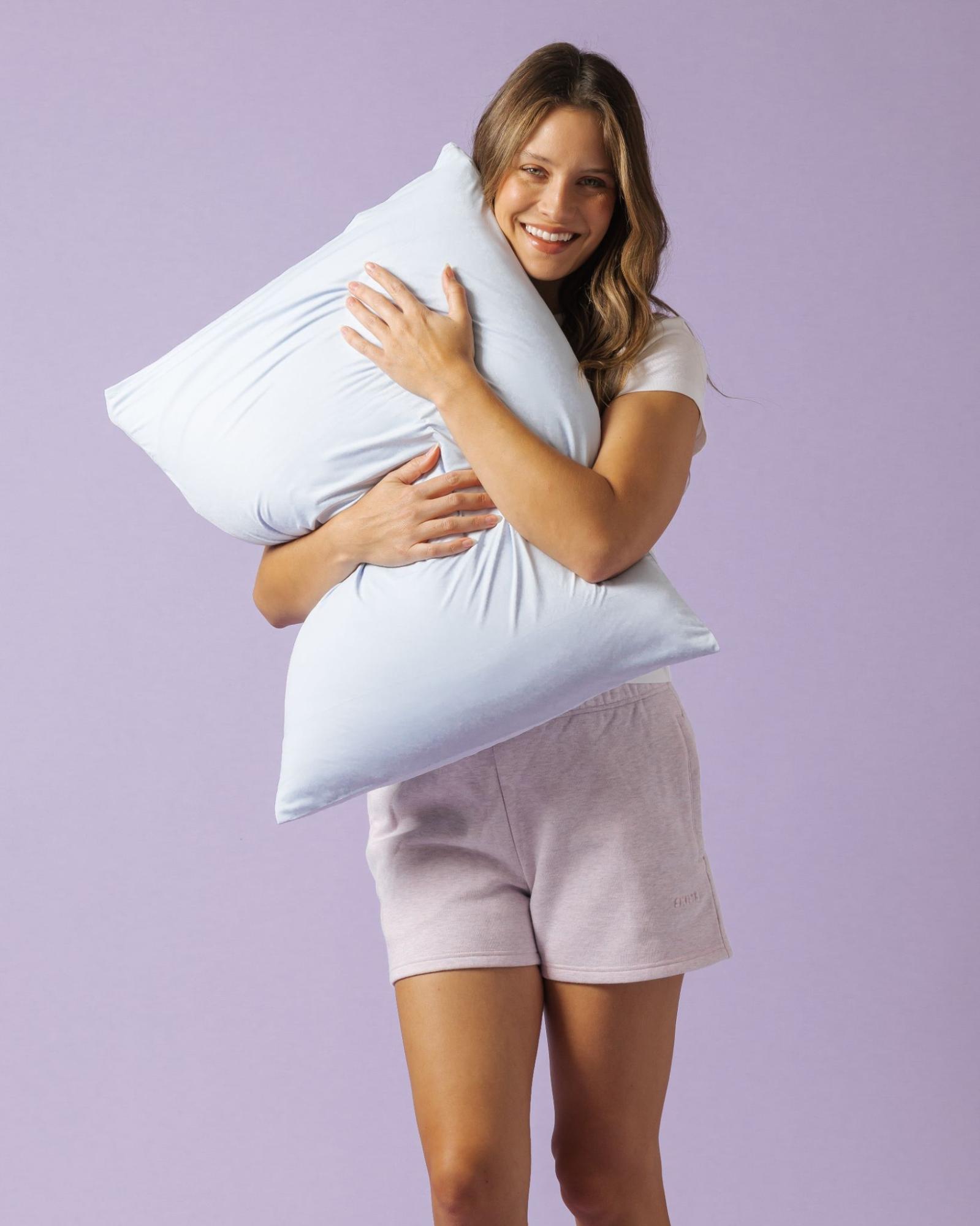 A smiling woman in a white t-shirt and shorts hugs a large, light blue pillow.