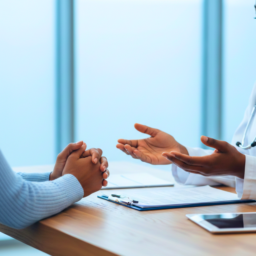 A doctor in a white coat gestures while speaking with a patient across a desk.
