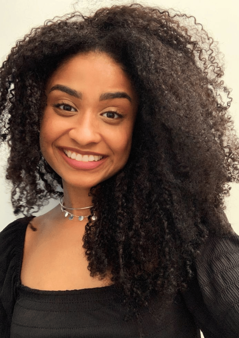 Smiling person with curly hair wearing a black top and necklace.
