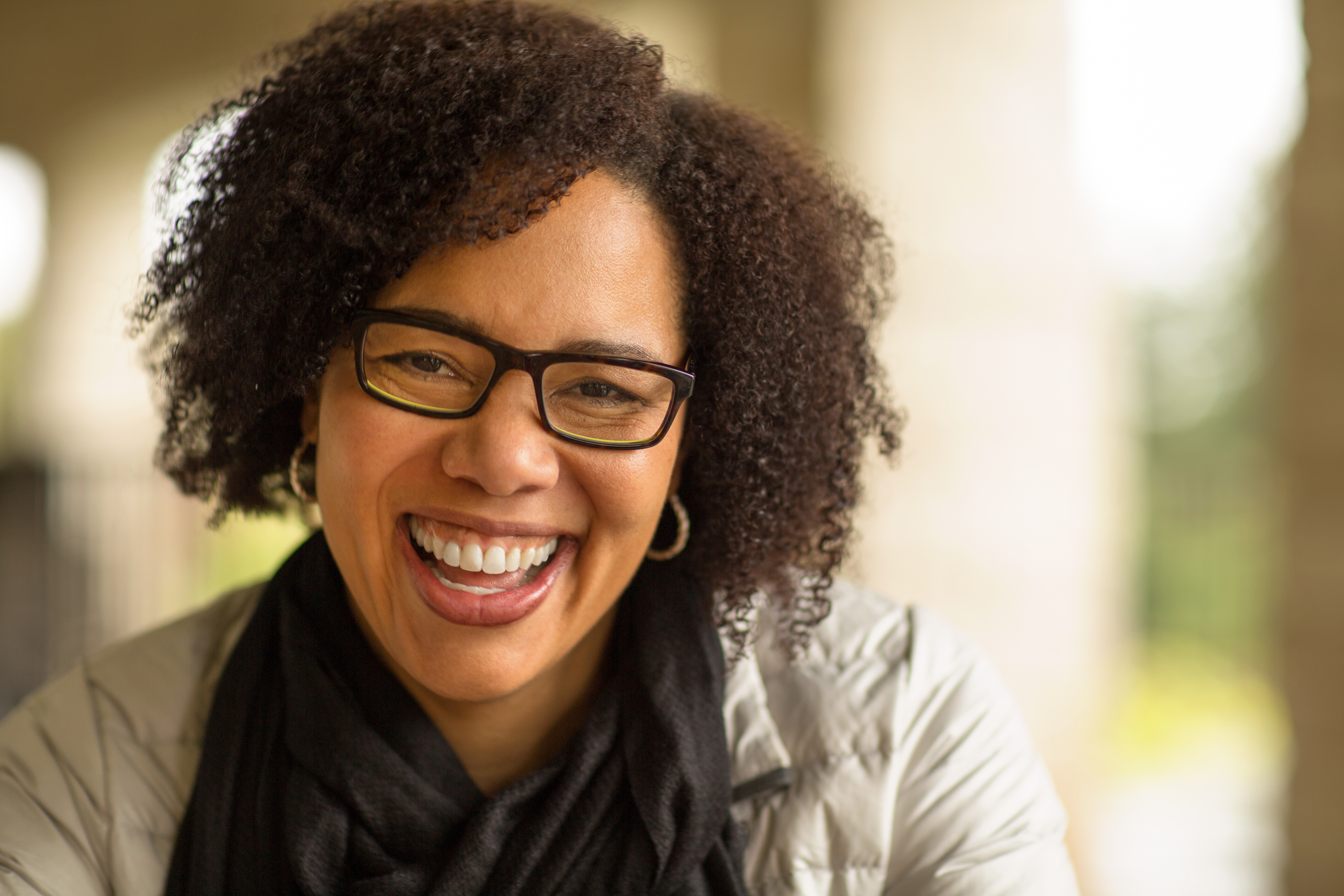 Smiling person with glasses and curly hair, wearing a scarf and jacket.