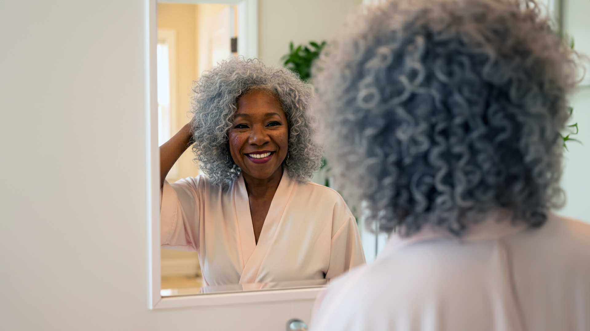 An older Black woman with curly gray hair in a pink robe smiles at her reflection.