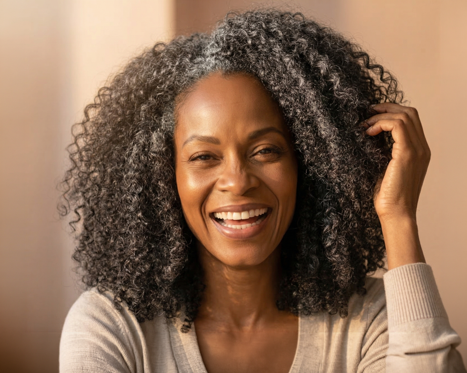 A smiling Black woman with curly, salt-and-pepper hair touches her hair while looking at the camera.