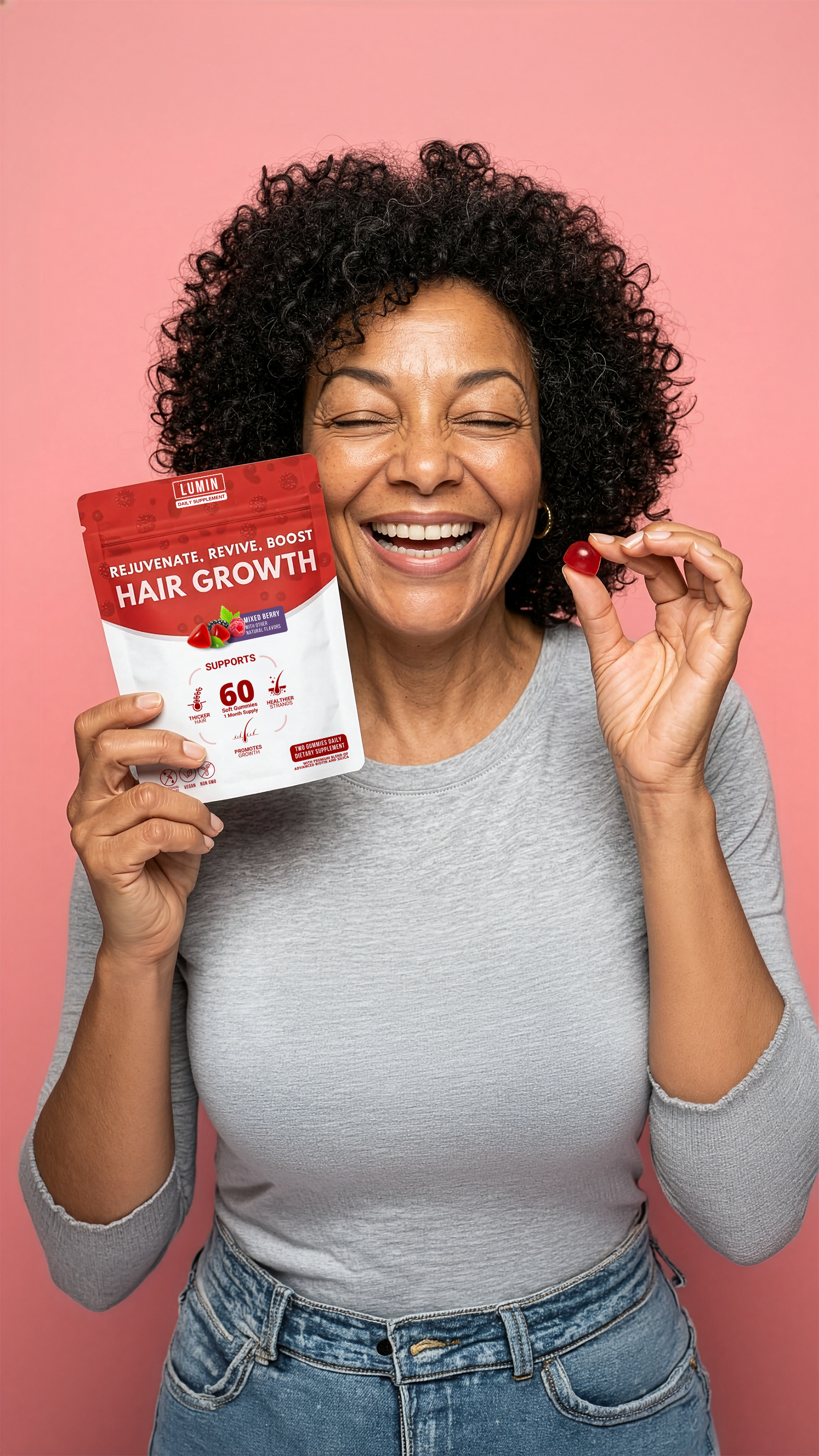 A smiling woman with her eyes closed holds a pouch of hair growth supplements and one red gummy.