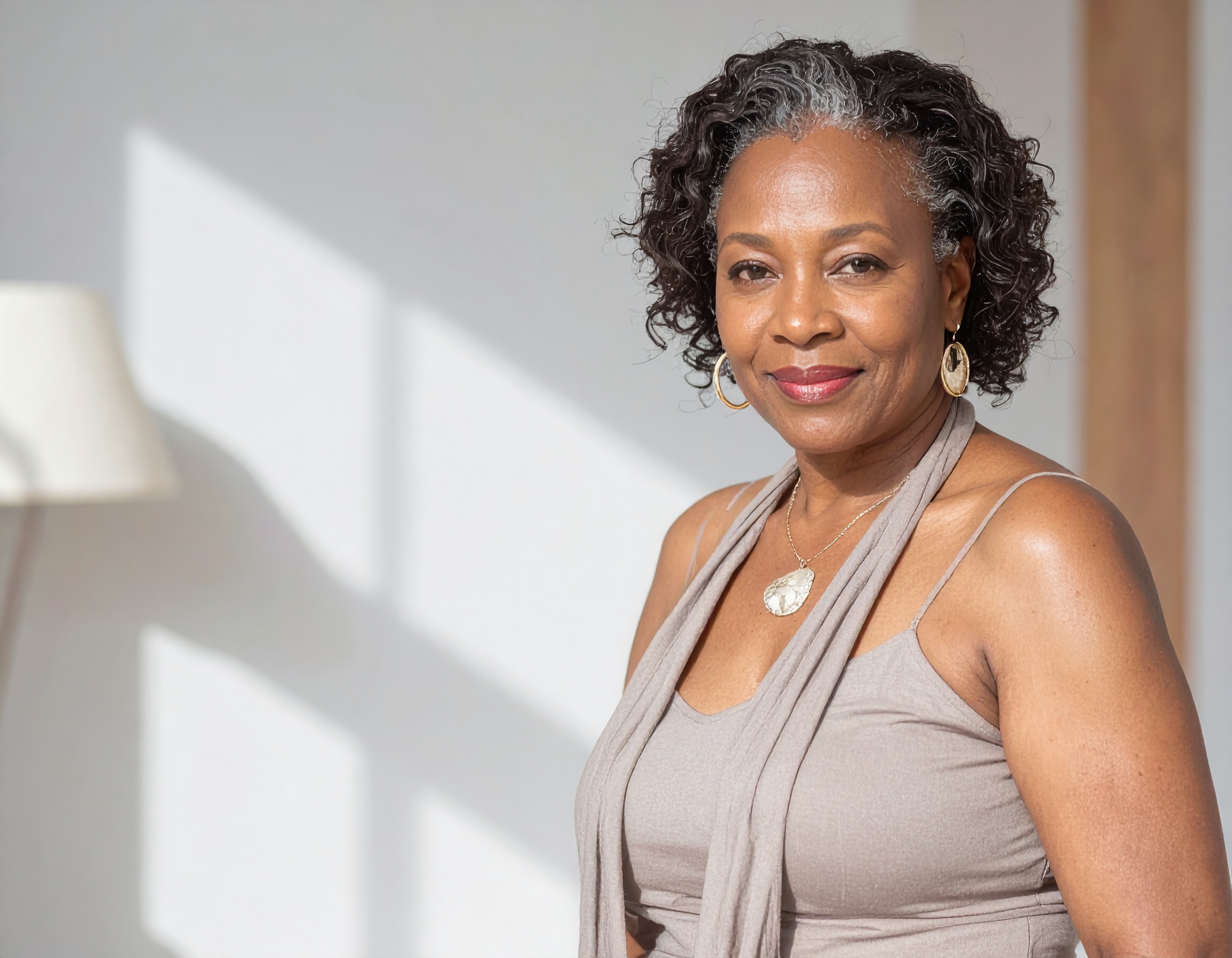 Smiling woman with short curly hair wearing a sleeveless top and earrings, standing in sunlight.