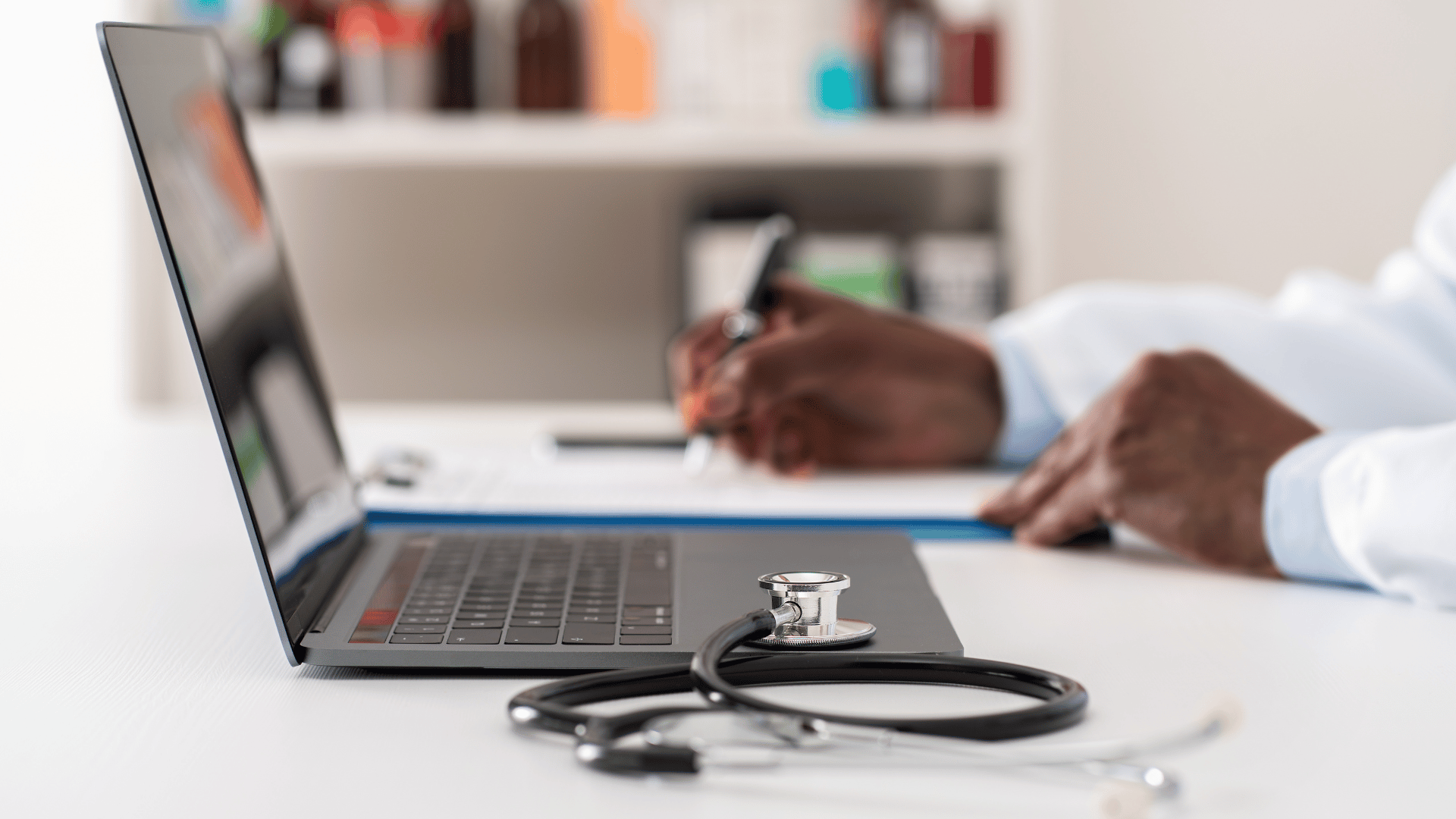 Stethoscope on desk beside laptop, person writing in background.