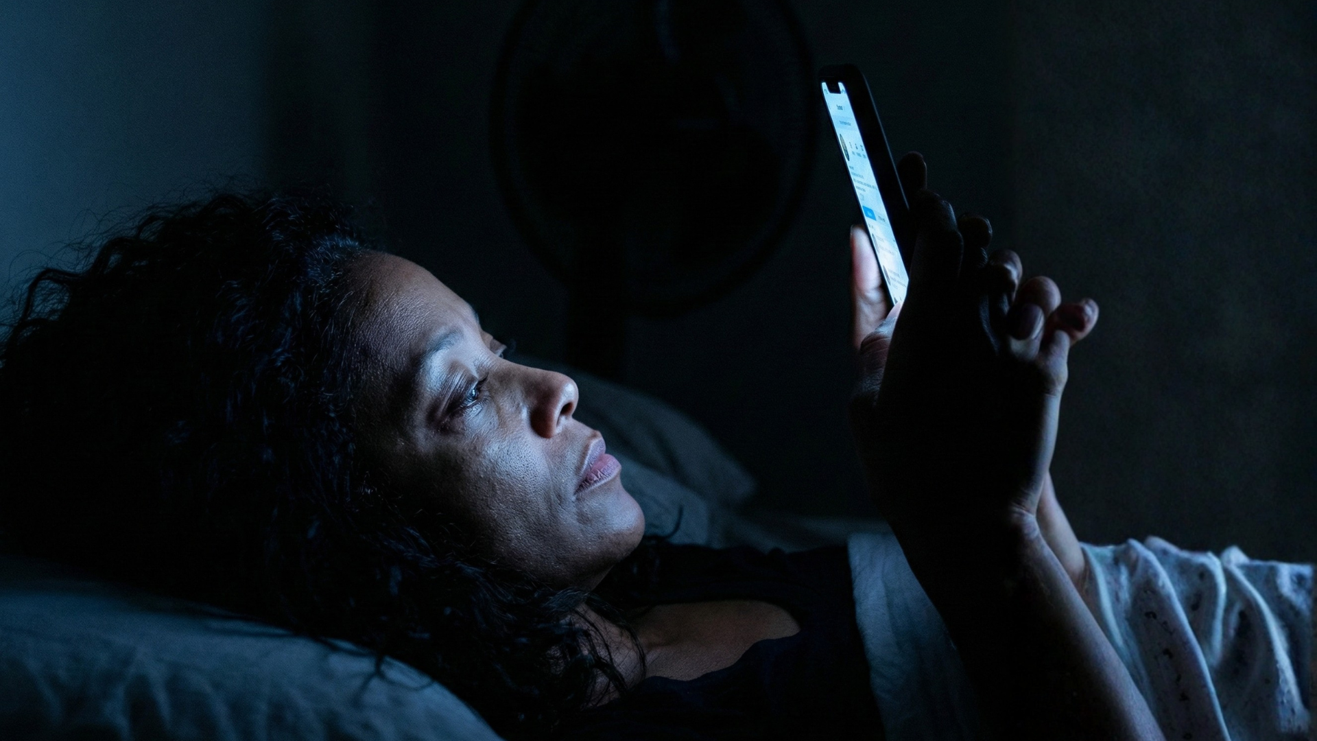 A woman lying in bed in the dark, her face illuminated by the light from her smartphone.