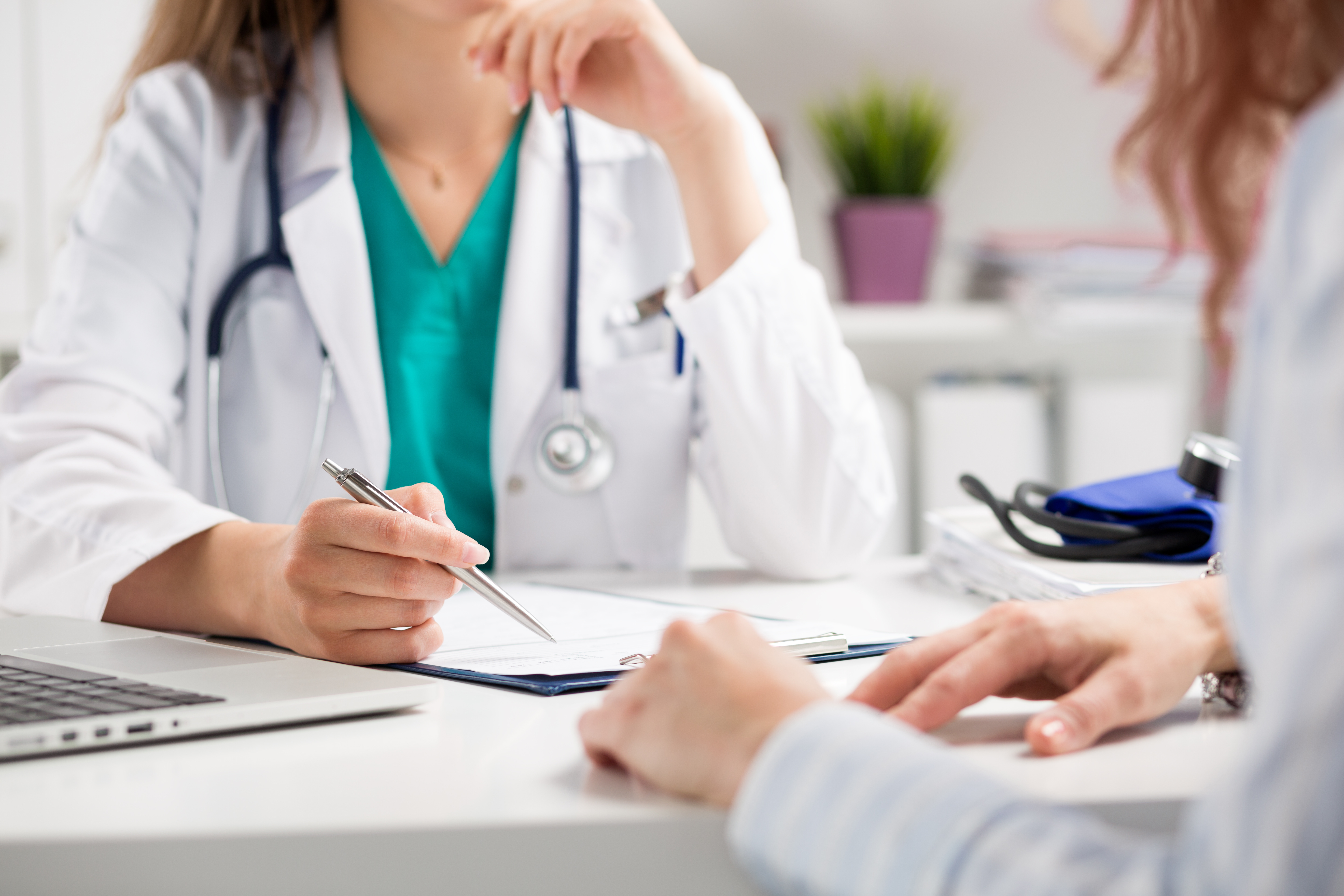 Doctor consulting with a patient, holding a pen and clipboard, stethoscope visible.