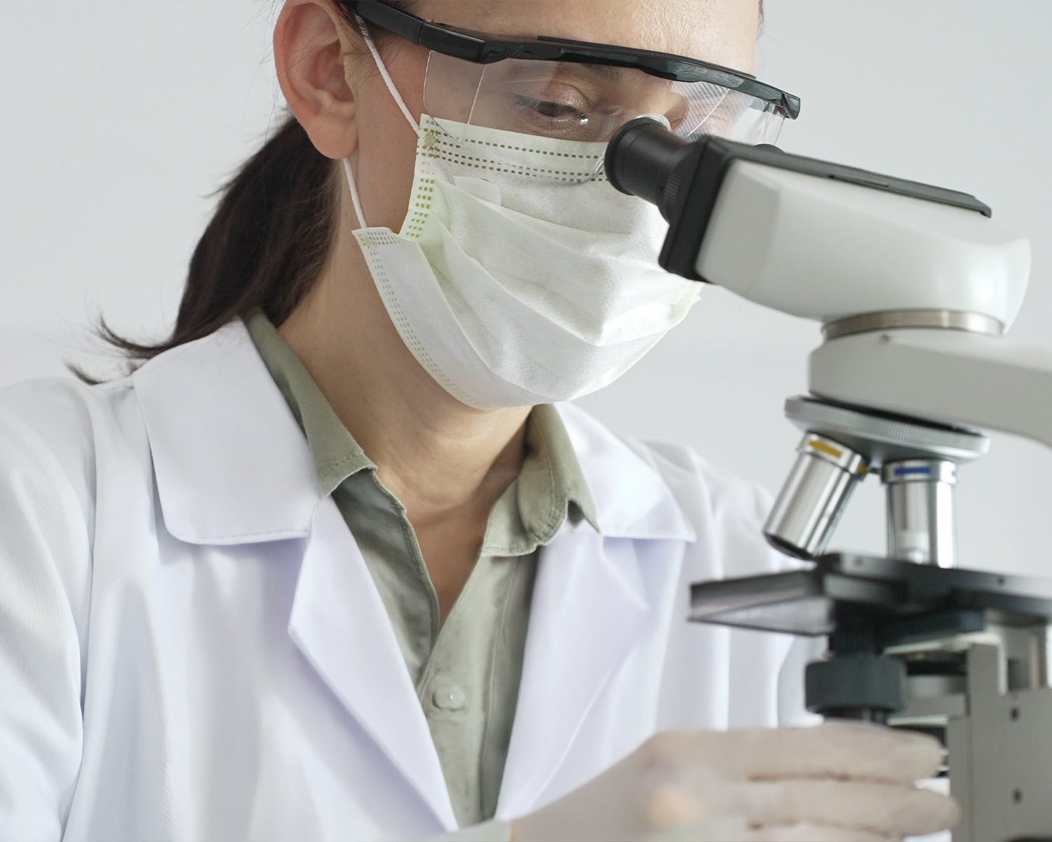 A scientist wearing a lab coat, face mask, and safety goggles looks into a microscope.