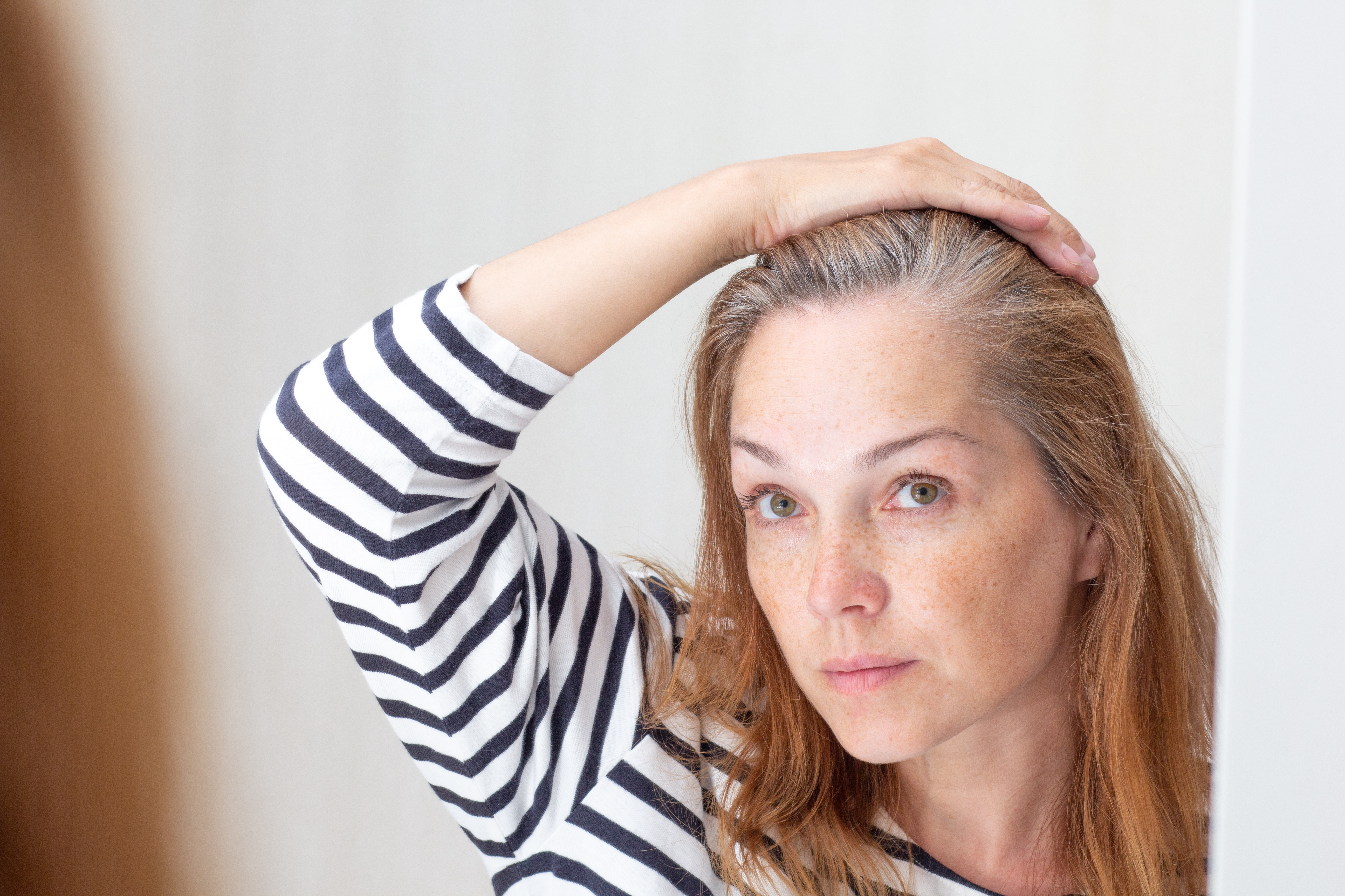 Woman with striped shirt looking in mirror, touching her hair.